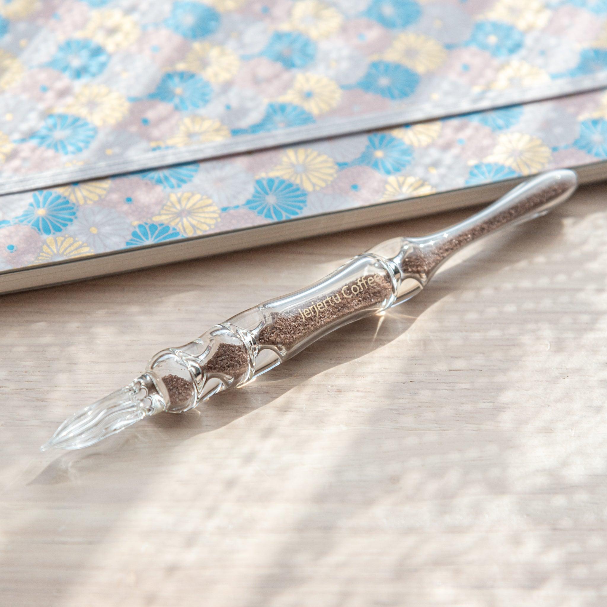 A glass dip pen with brown sand-like particles inside its shaft, branded with "Jerjertu coffee," is placed on a wooden surface near a pastel floral notebook.