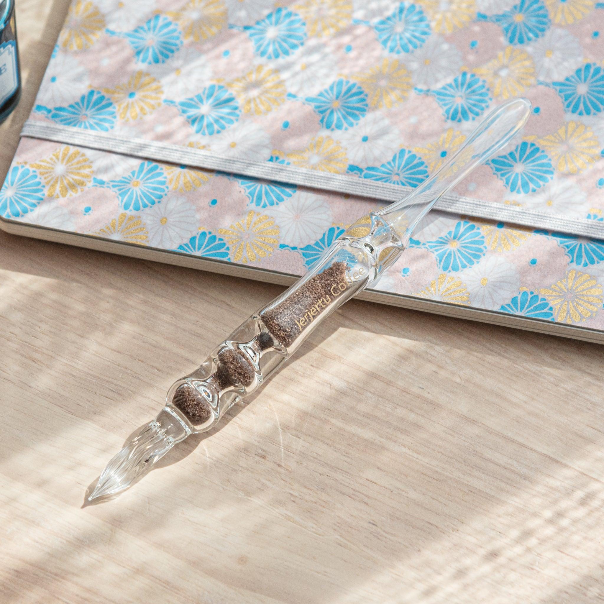 A glass dip pen with brown sand-like particles inside its shaft, branded with "Jerjertu coffee," is placed on a wooden surface near a pastel floral notebook.