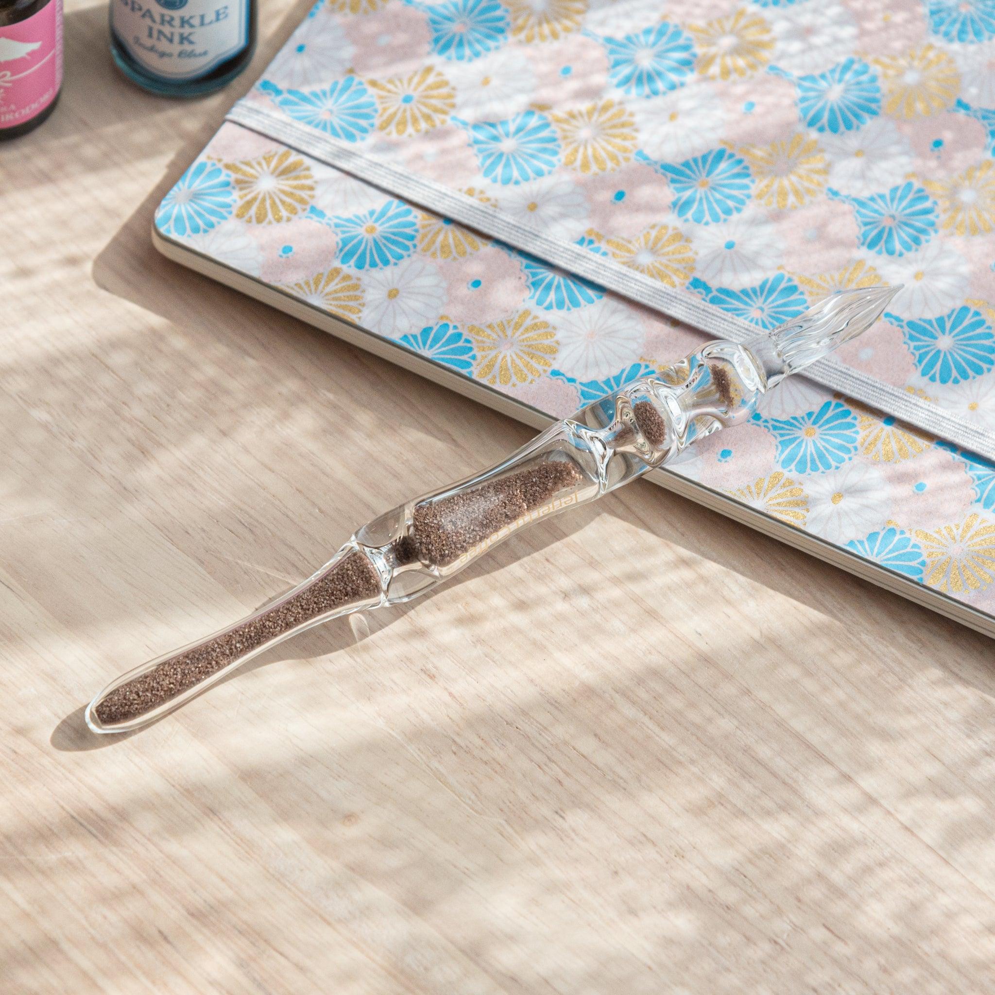 A glass dip pen with brown sand-like particles inside its shaft, branded with "Jerjertu coffee," is placed on a wooden surface near a pastel floral notebook.