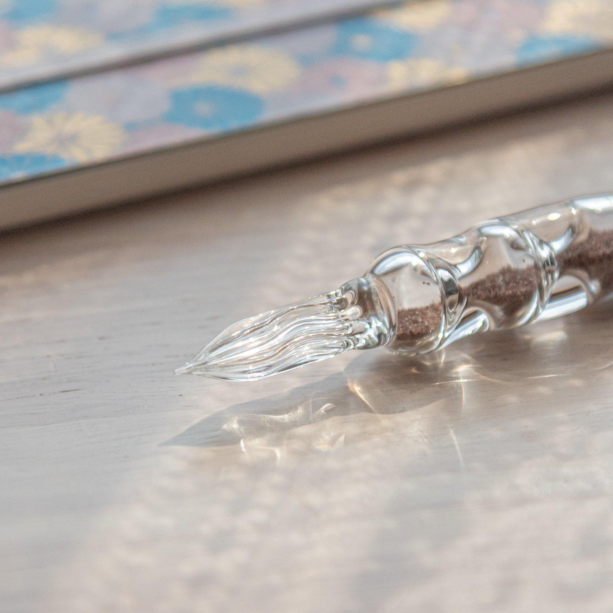 A glass dip pen with brown sand-like particles inside its shaft, branded with "Jerjertu coffee," is placed on a wooden surface near a pastel floral notebook.