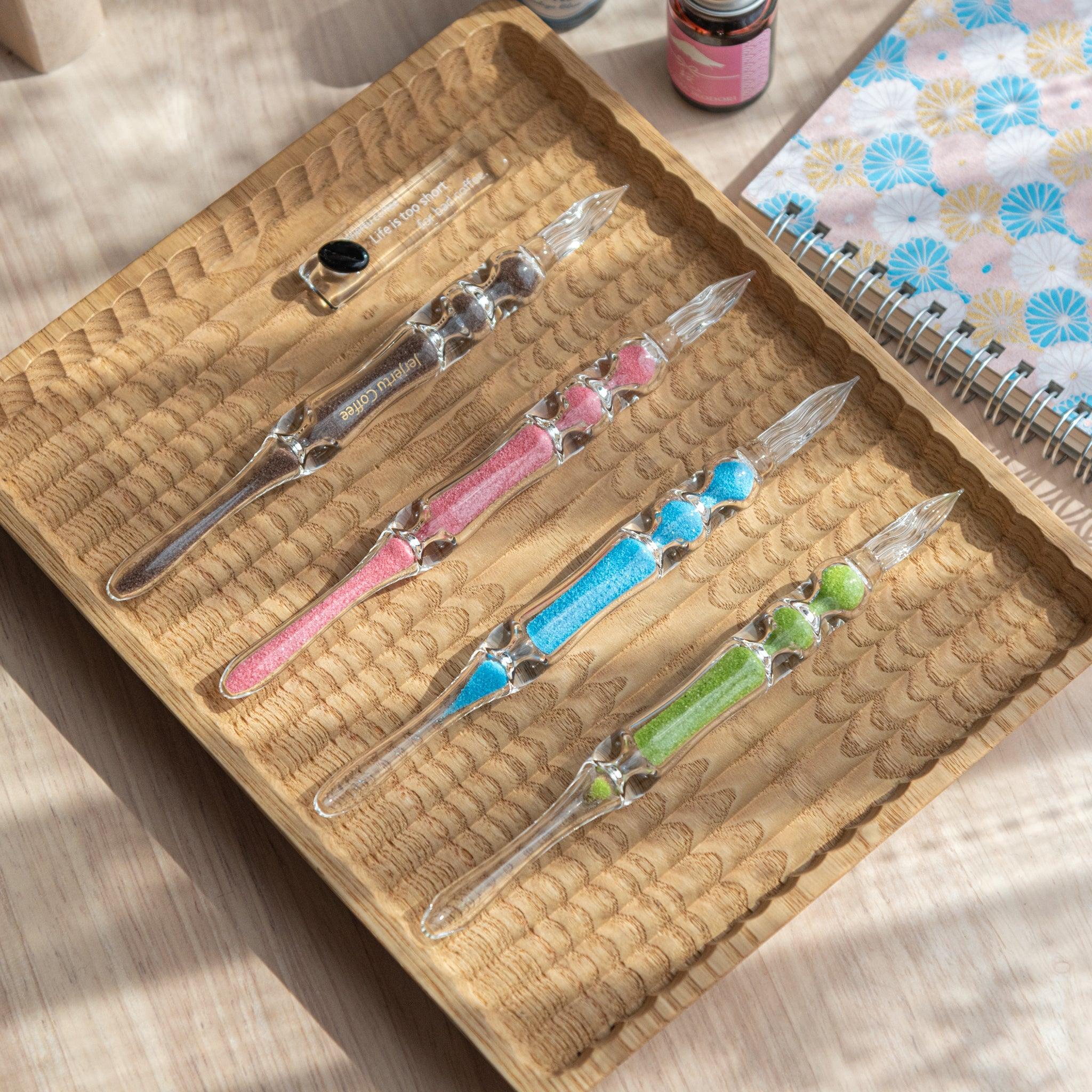 A set of four glass dip pens with sand-like particles in brown, pink, blue, and green inside their shafts are neatly displayed on a carved wooden tray. A small pen holder with a black coffee bean design is placed alongside the pens.