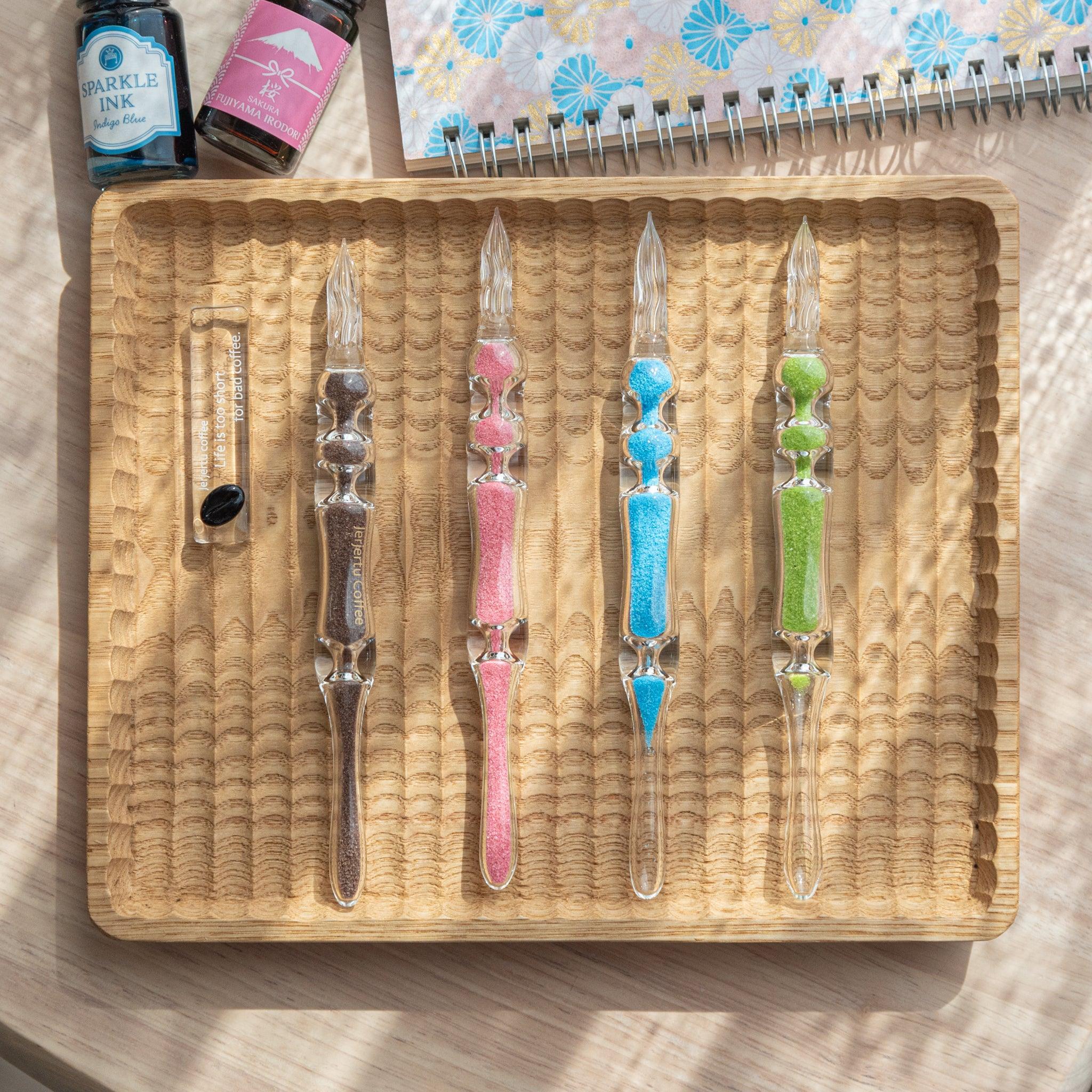 A set of four glass dip pens with brown, pink, blue, and green sand-like particles inside their shafts are arranged on a wooden tray. The pens are displayed next to a small glass pen holder with the words "Life is too short for bad coffee."