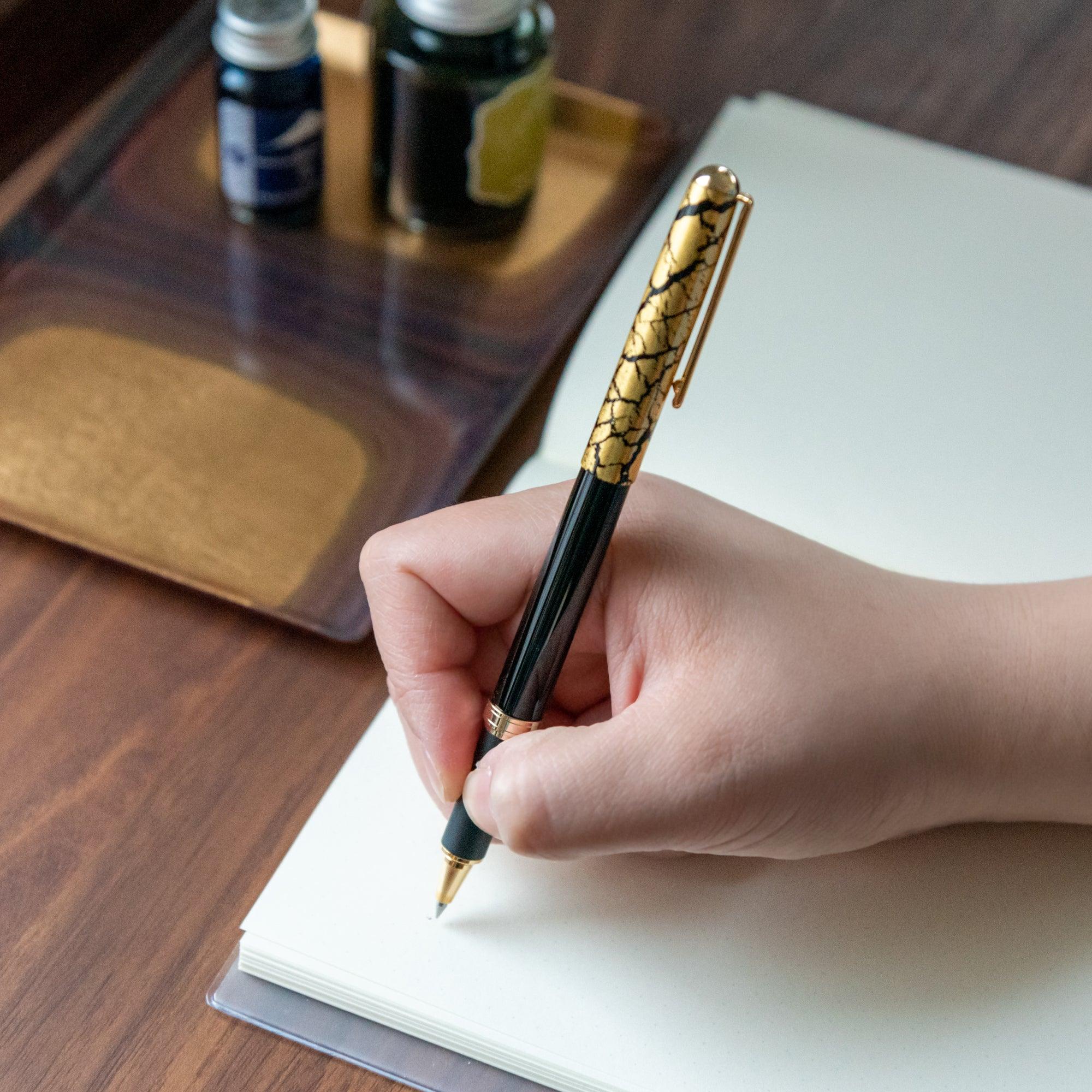 A person writing with a black pen featuring a gold crack pattern cap, with ink bottles and a wooden tray in the background.