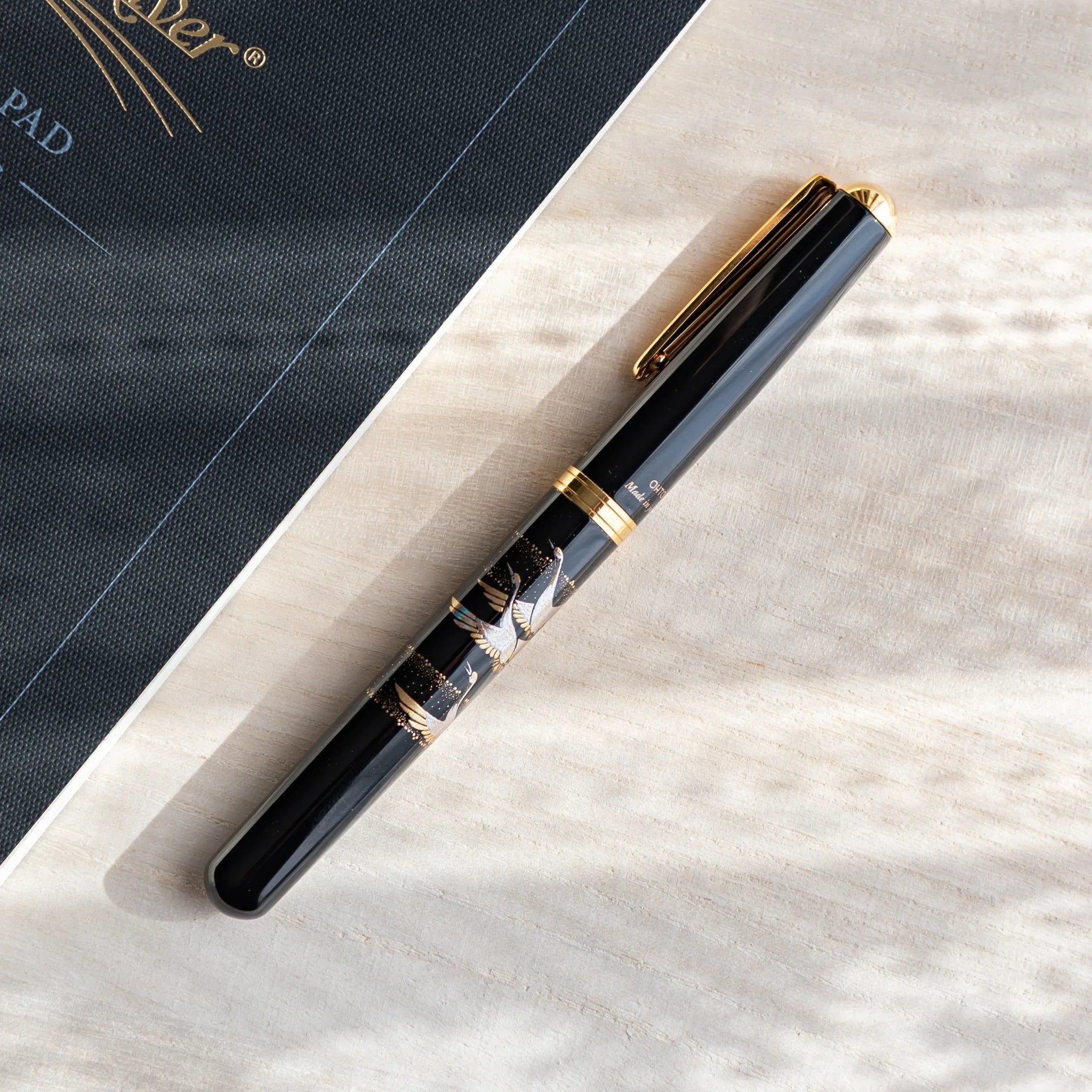 Black Yamanaka Japanese fountain pen with silver crane artwork resting diagonally on a light wooden desk beside a notebook.