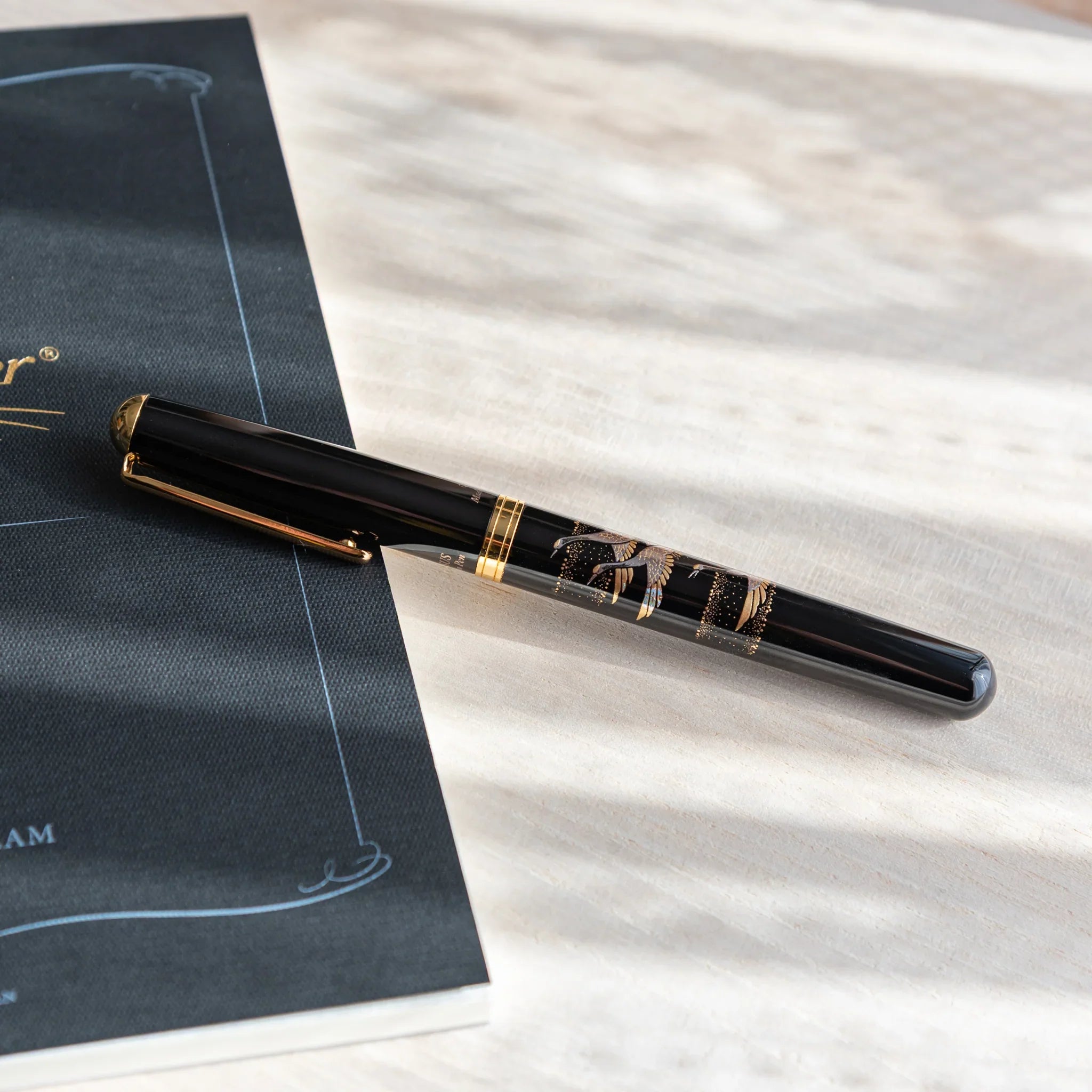 Black Yamanaka Japanese fountain pen with silver crane artwork resting on a light wooden desk beside a black notebook.