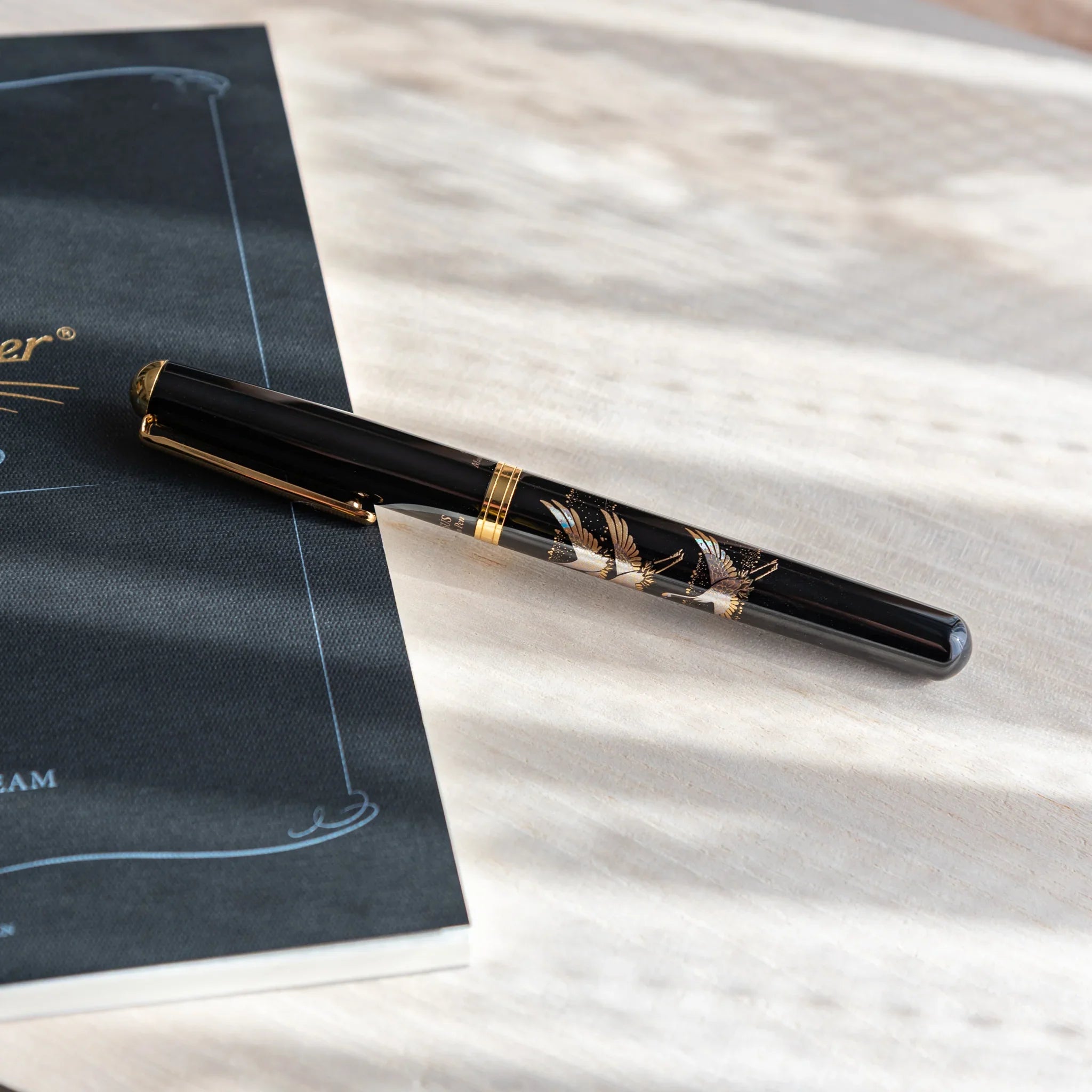 Black Japanese fountain pen with silver crane artwork resting diagonally on a light wood desk beside a dark notebook.
