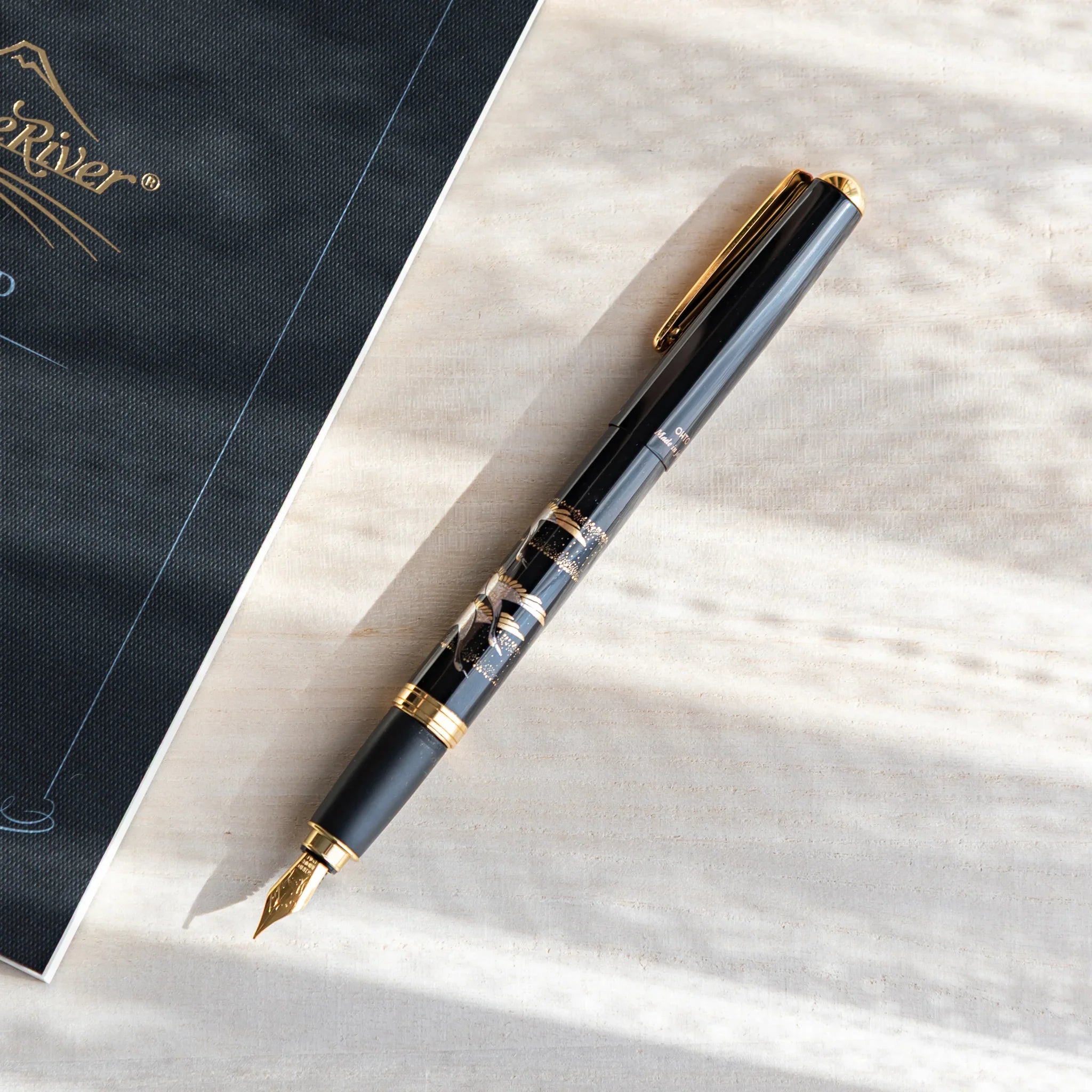 Black Japanese fountain pen with silver crane artwork and gold trim resting diagonally on a wood desk beside a dark notebook.
