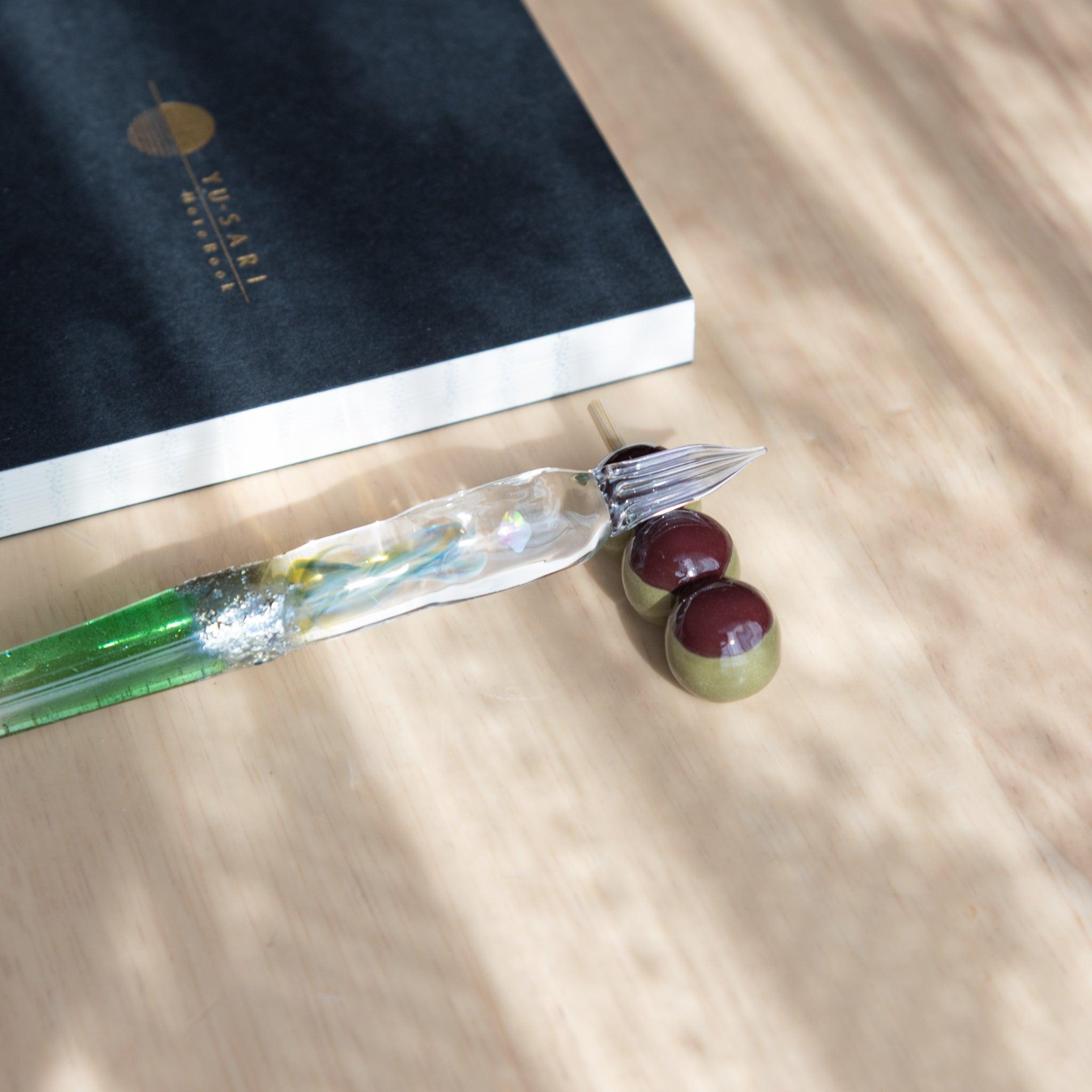 A handmade ceramic pen rest shaped as a mugwort dango skewer, holding a green glass dip pen. Showcasing Japanese stationery artistry.