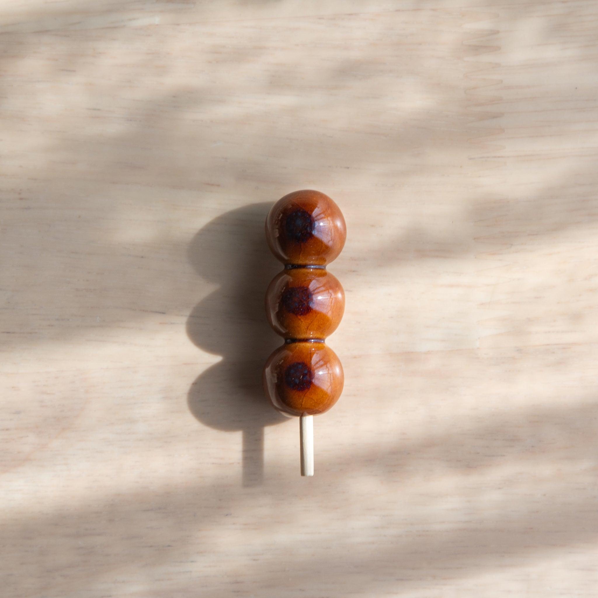 A handmade ceramic pen rest shaped as a mitarashi dango skewer, holding a green and blue glass dip pen. Part of traditional Japanese stationery items.