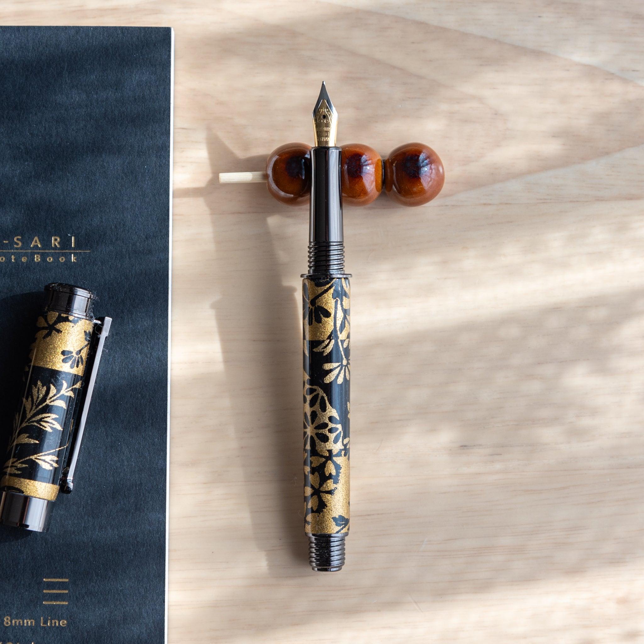 A handmade ceramic pen rest shaped as a mugwort dango skewer, holding a black and gold fountain pen. Part of traditional Japanese stationery items.