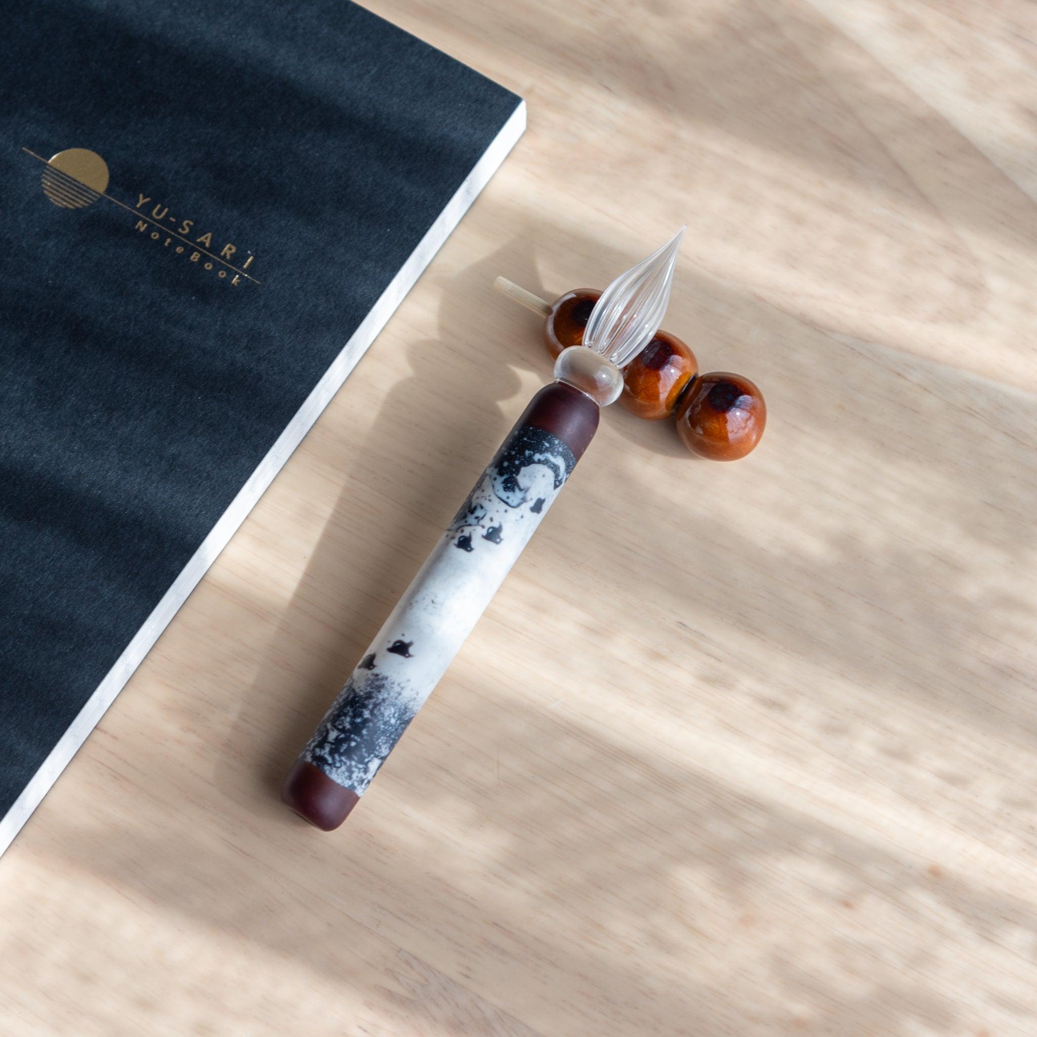 A handmade ceramic pen rest shaped as a mitarashi dango skewer, holding a black and white glass dip pen. Showcasing Japanese stationery artistry.