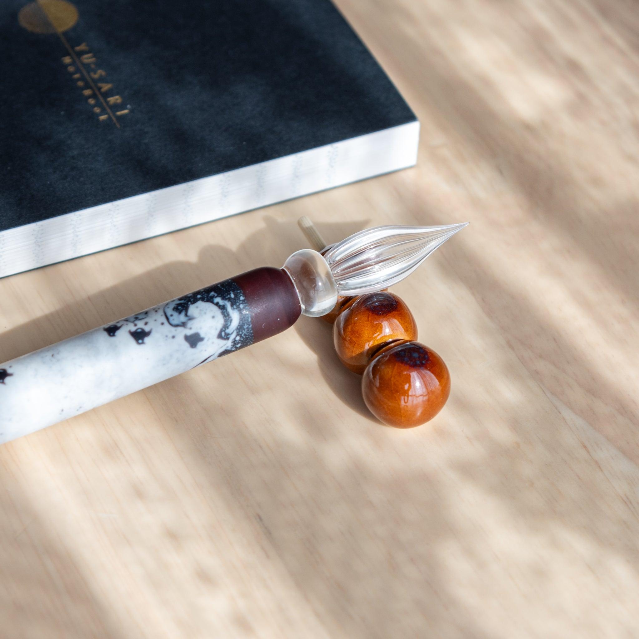 A handmade ceramic pen rest shaped as a mitarashi dango skewer, holding a black and white glass dip pen. Showcasing Japanese stationery artistry.