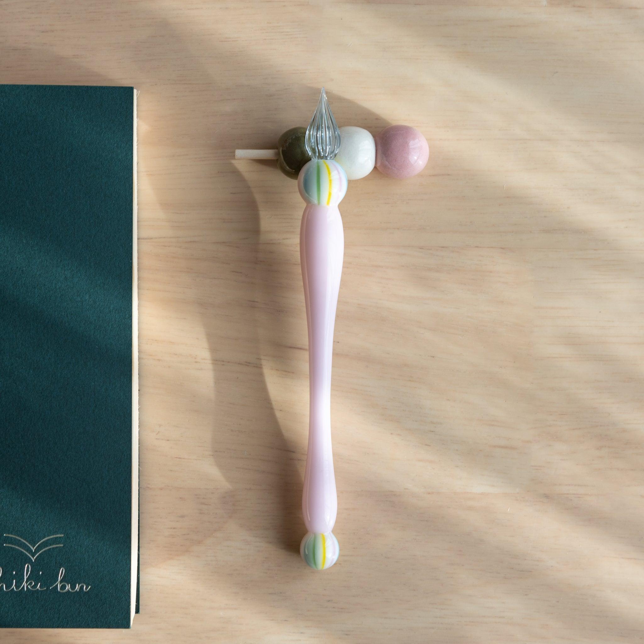 A handmade ceramic pen rest shaped as a hanami dango skewer, holding a pink glass dip pen. Part of traditional Japanese stationery items.