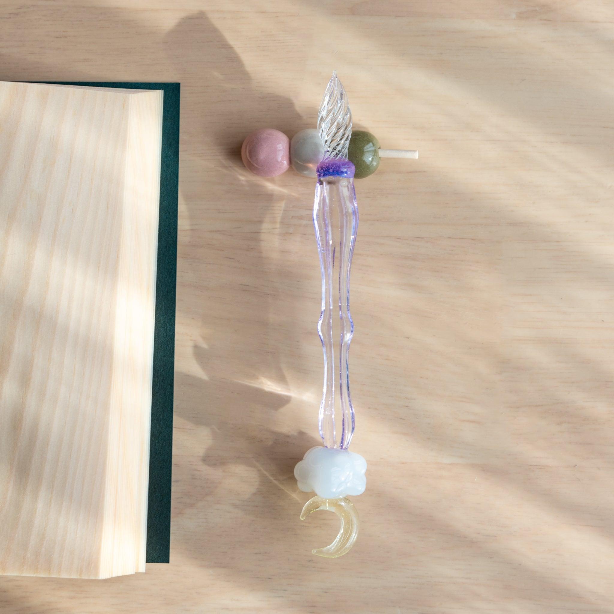 A handmade ceramic pen rest shaped as a hanami dango skewer, holding a purple glass dip pen with a moon and cloud motif. Japanese stationery item.