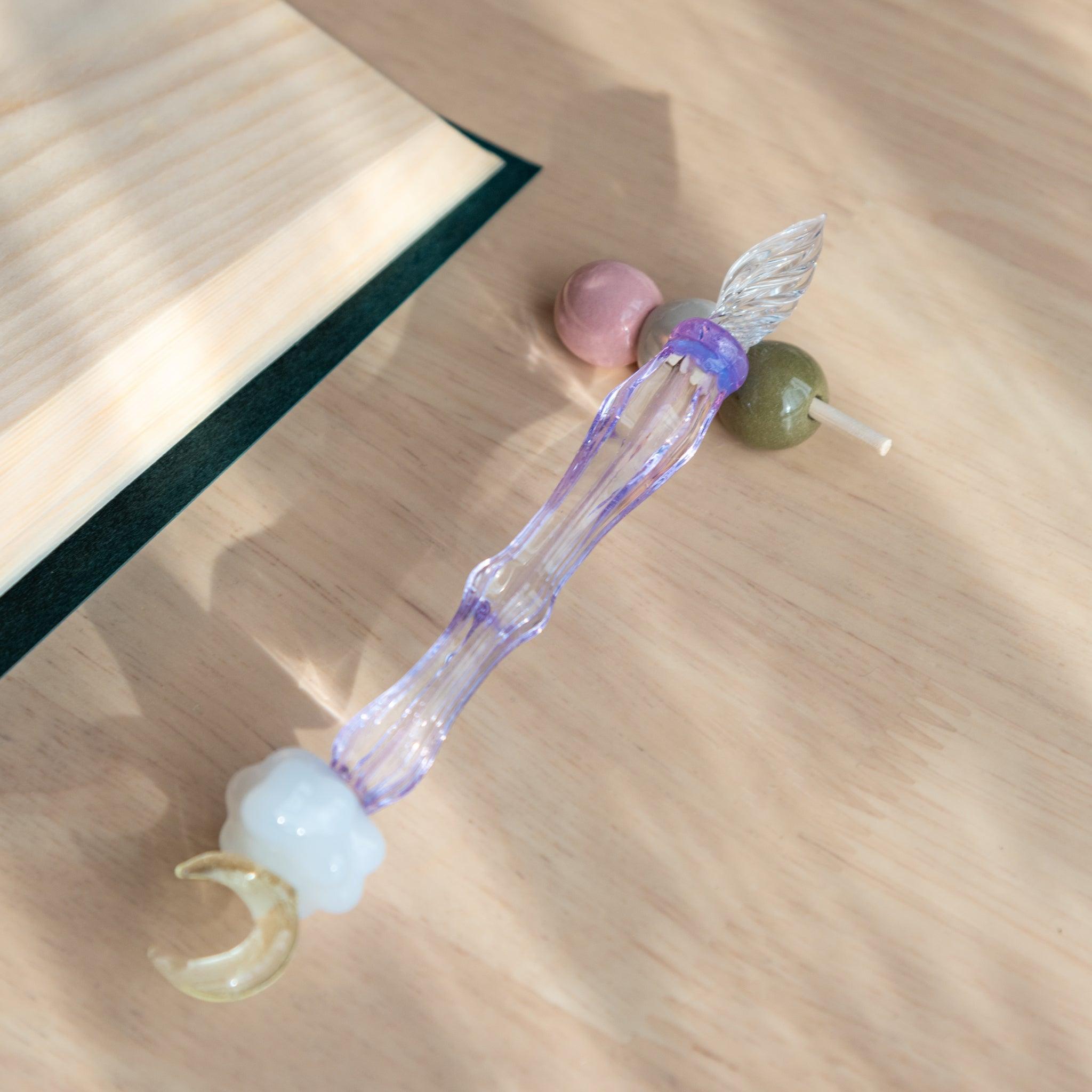 A handmade ceramic pen rest shaped as a hanami dango skewer, holding a purple glass dip pen with a moon and cloud motif. Japanese stationery item.