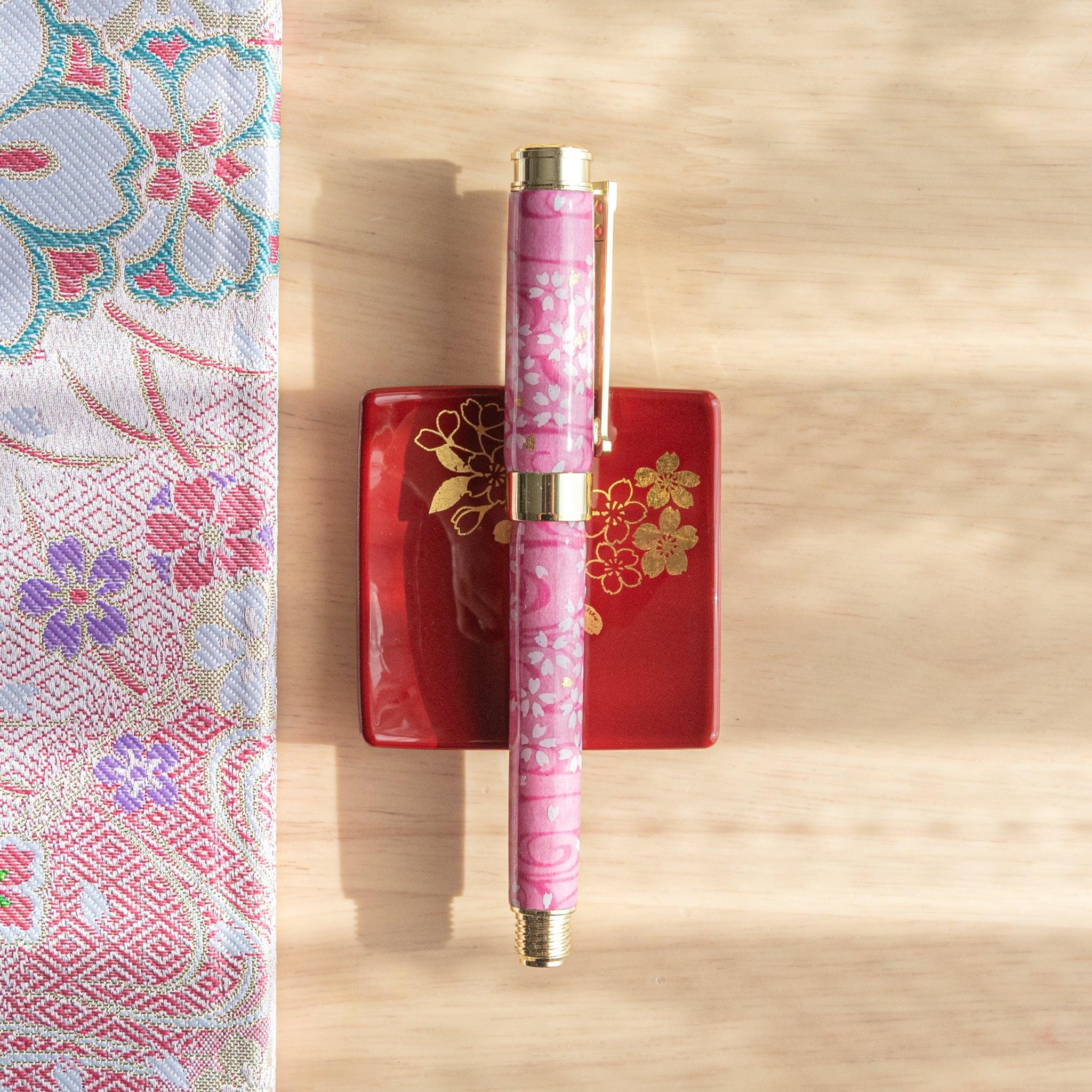 Pink and gold fountain pen with sakura motif on red tray and patterned fabric in soft sunlight.