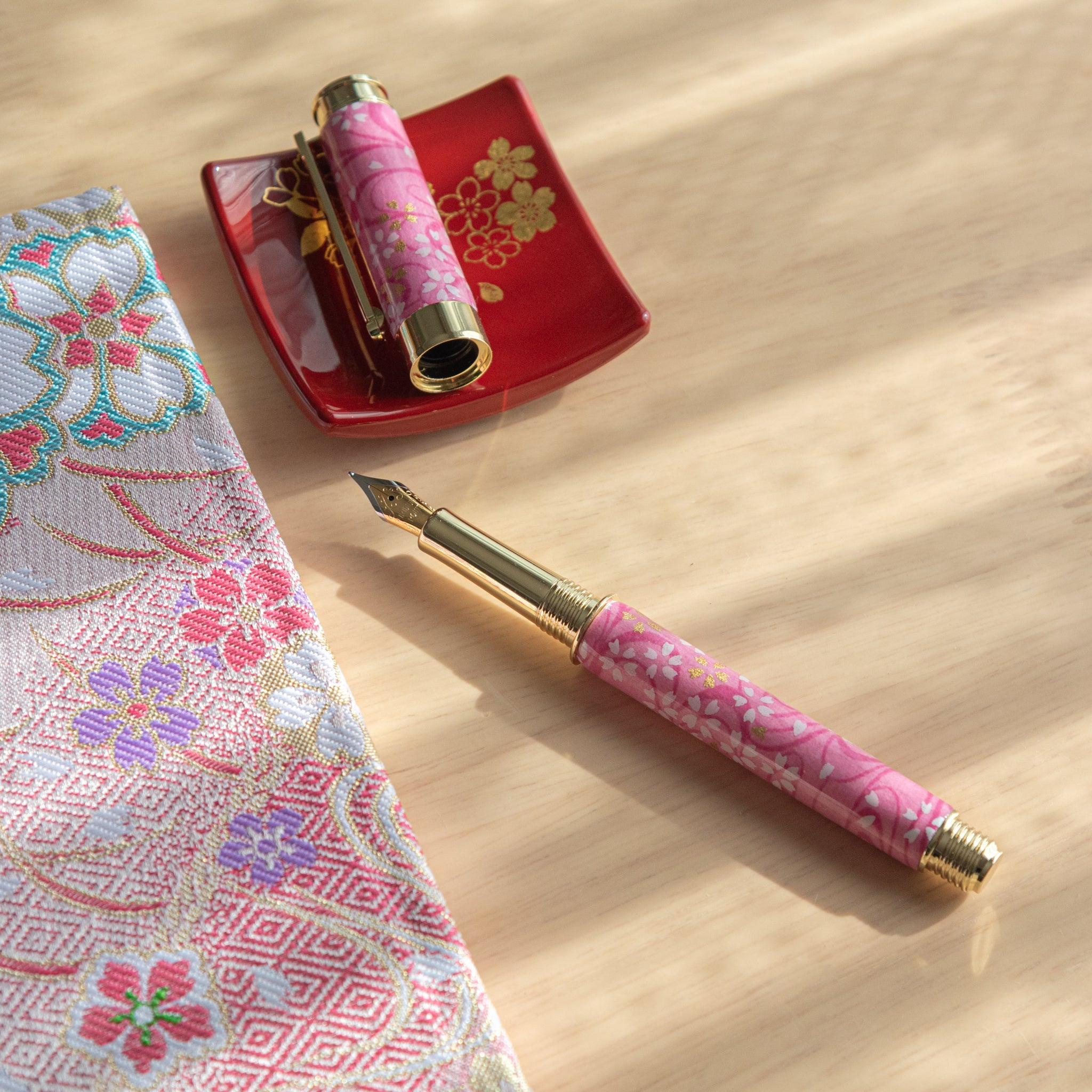 Open pink sakura-patterned fountain pen with gold accents on red tray, next to floral patterned fabric in natural light. (125 characters)