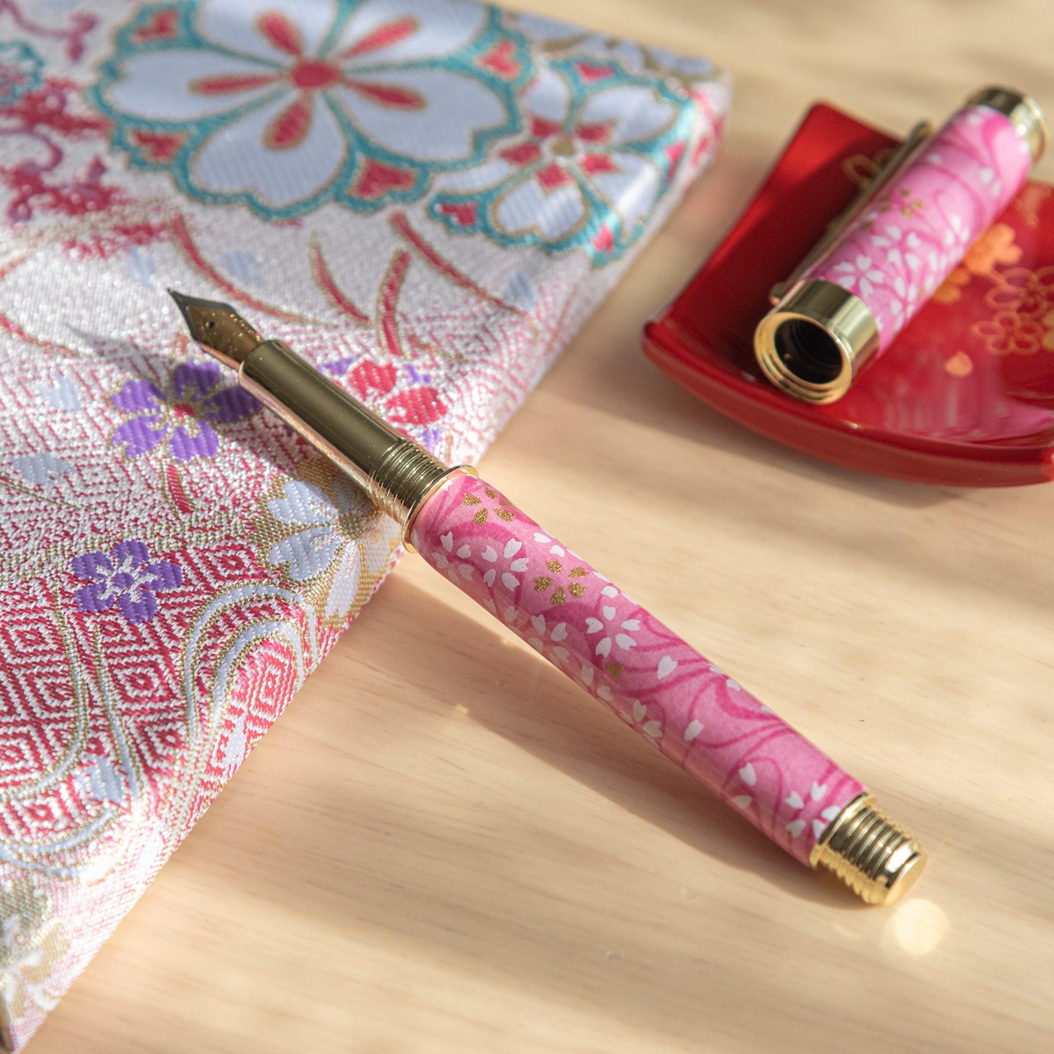 Sakura-patterned pink fountain pen with gold trim, open next to floral fabric and a red tray with golden floral design. (124 characters)