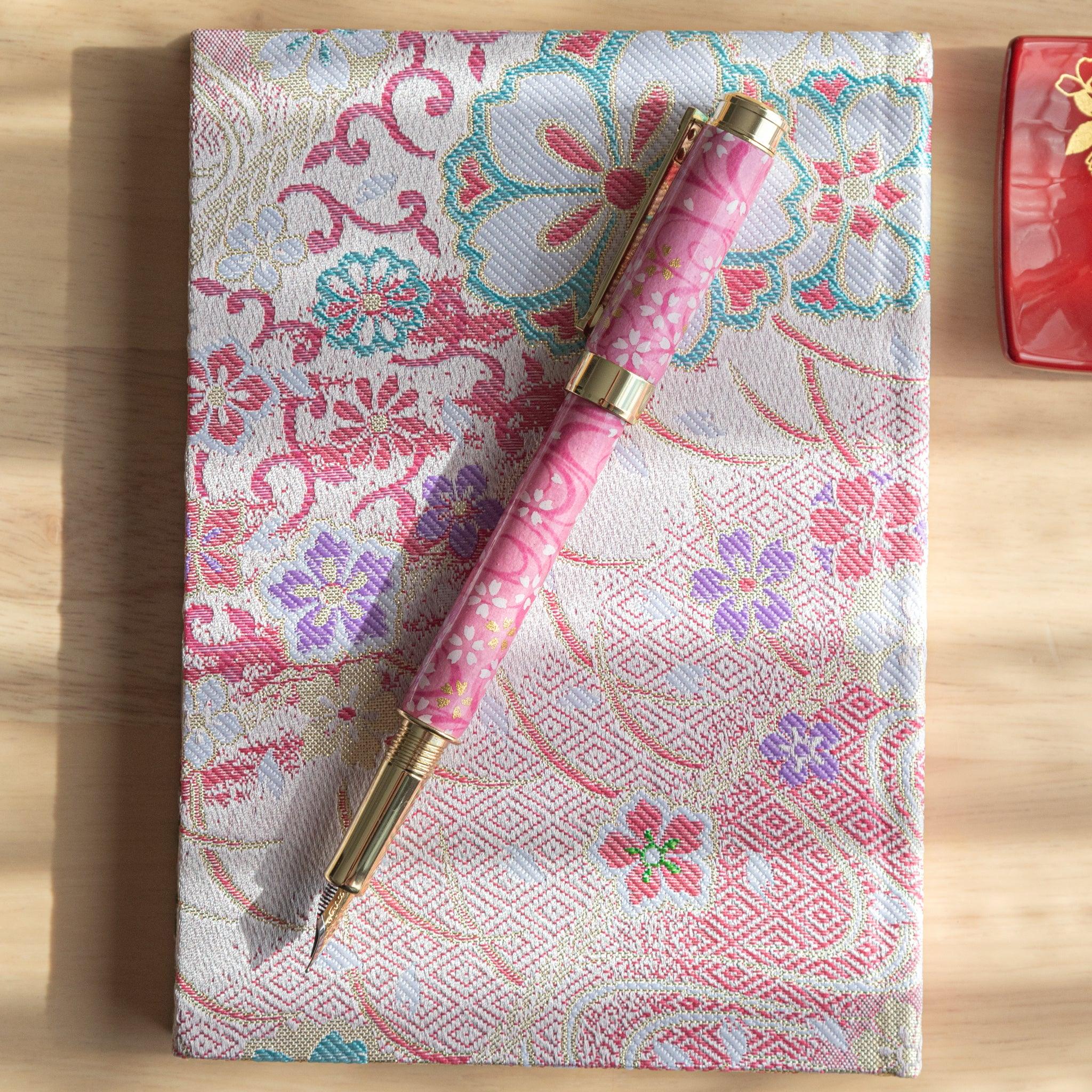 Pink sakura-themed fountain pen with gold accents placed on intricate floral fabric under soft sunlight. (114 characters)