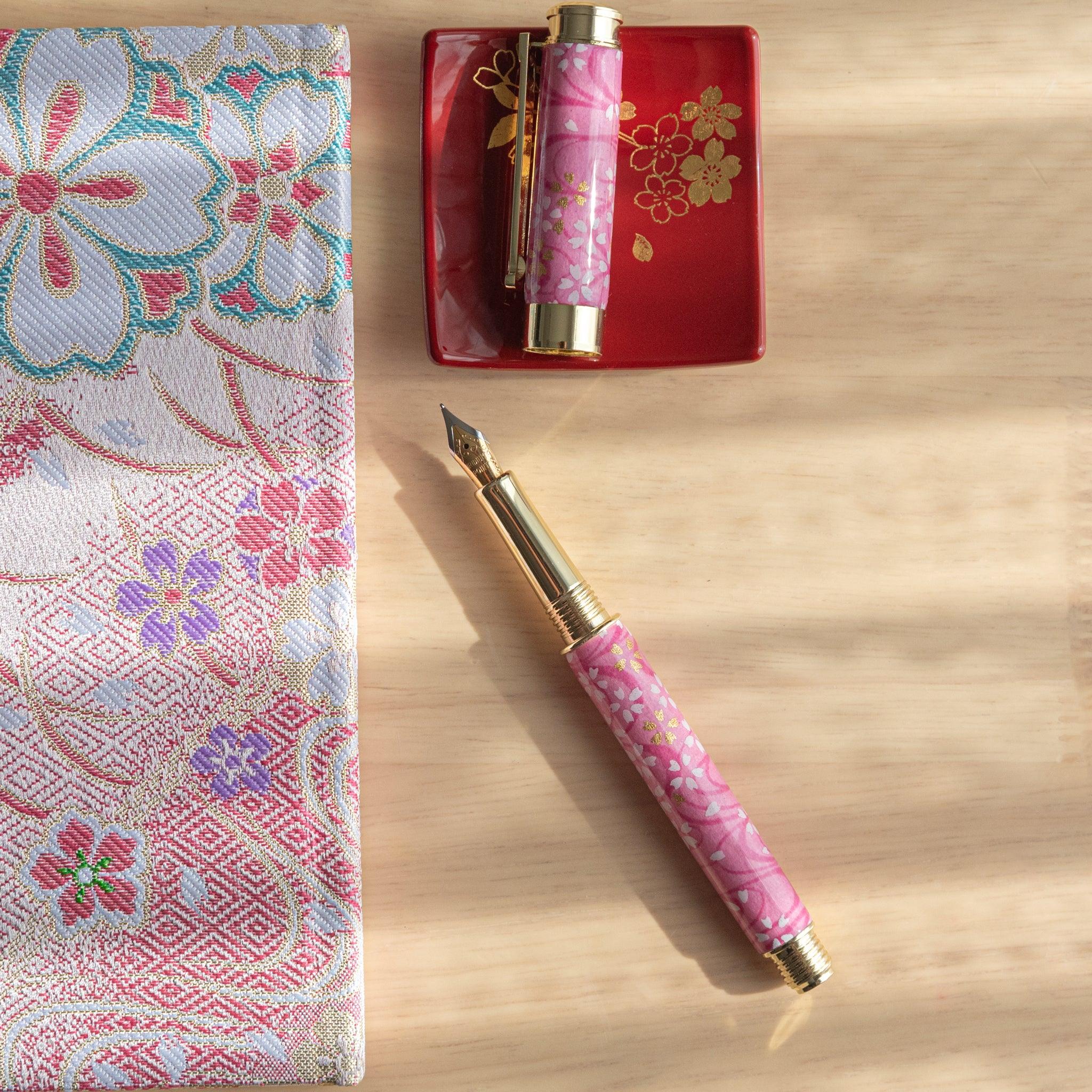 Pink sakura fountain pen with gold details, open on a red tray and floral fabric in sunlight. (99 characters)