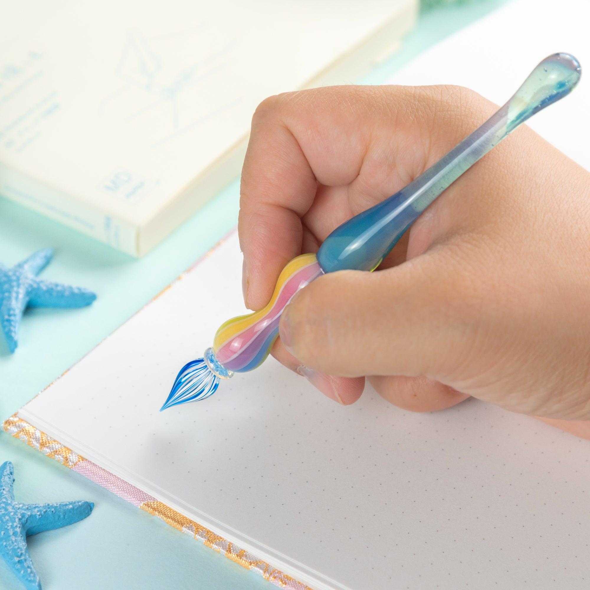 Close-up of a rainbow-colored glass pen in hand, writing in a notebook. A must-have for those who appreciate Japanese stationery and fine glass pens.