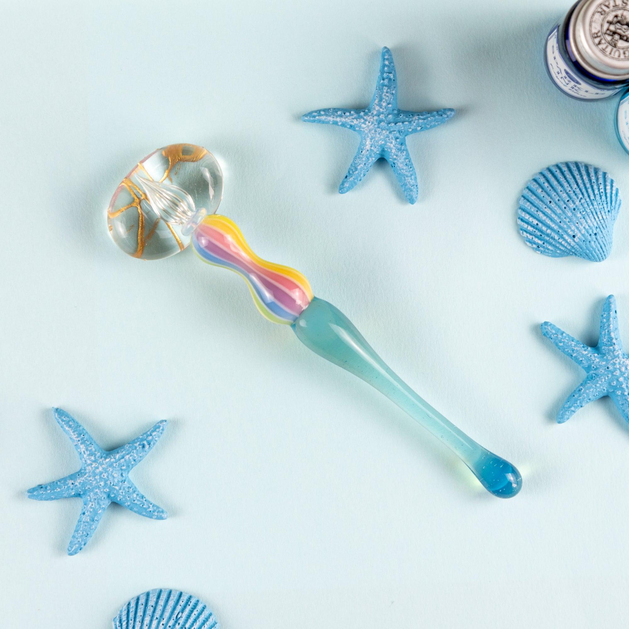 Rainbow balloon glass pen resting on a clear pen rest, surrounded by blue starfish and seashells. Perfect for Japanese stationery lovers.