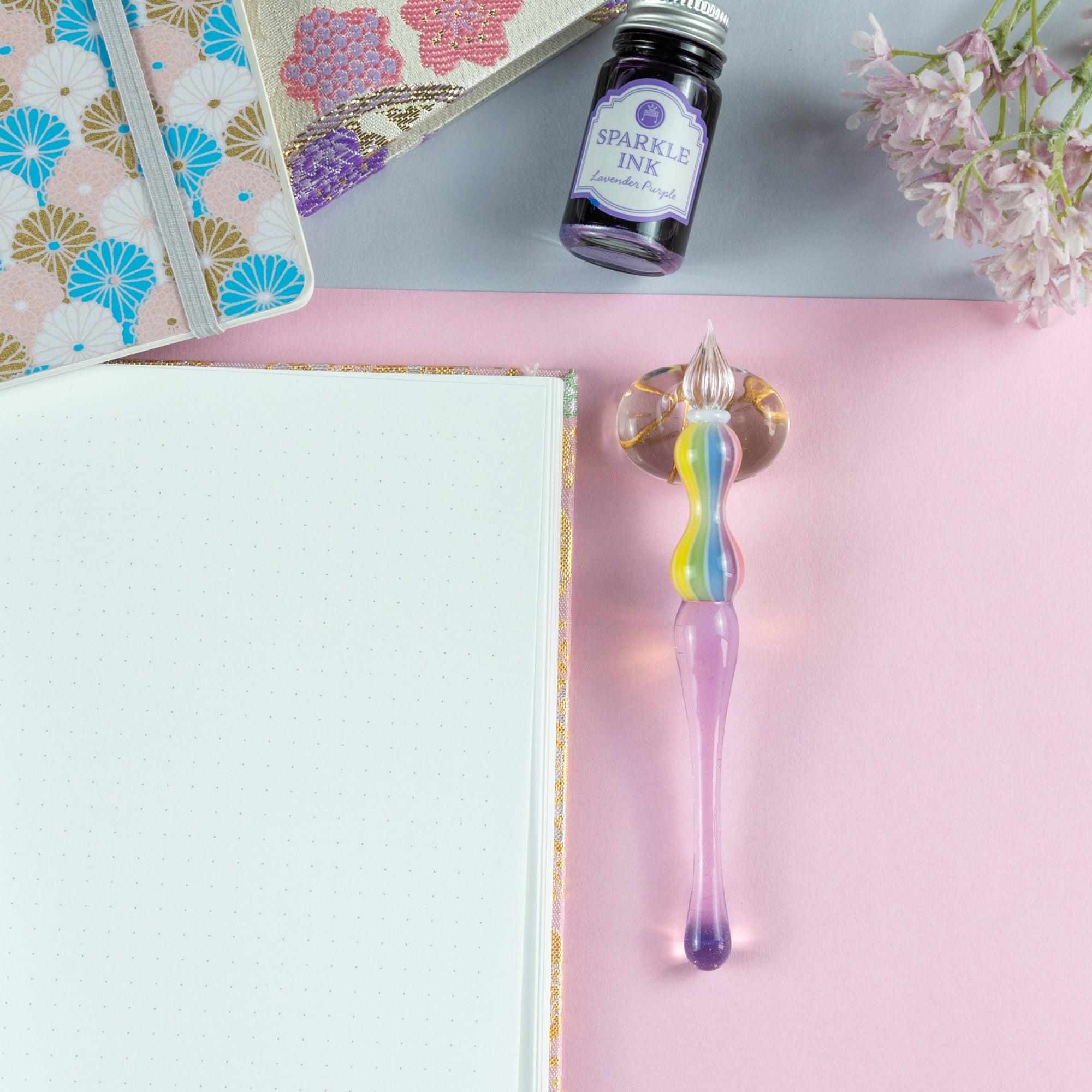 Glass pen with rainbow-colored handle resting on a floral-patterned notebook. Perfect for Japanese stationery enthusiasts.