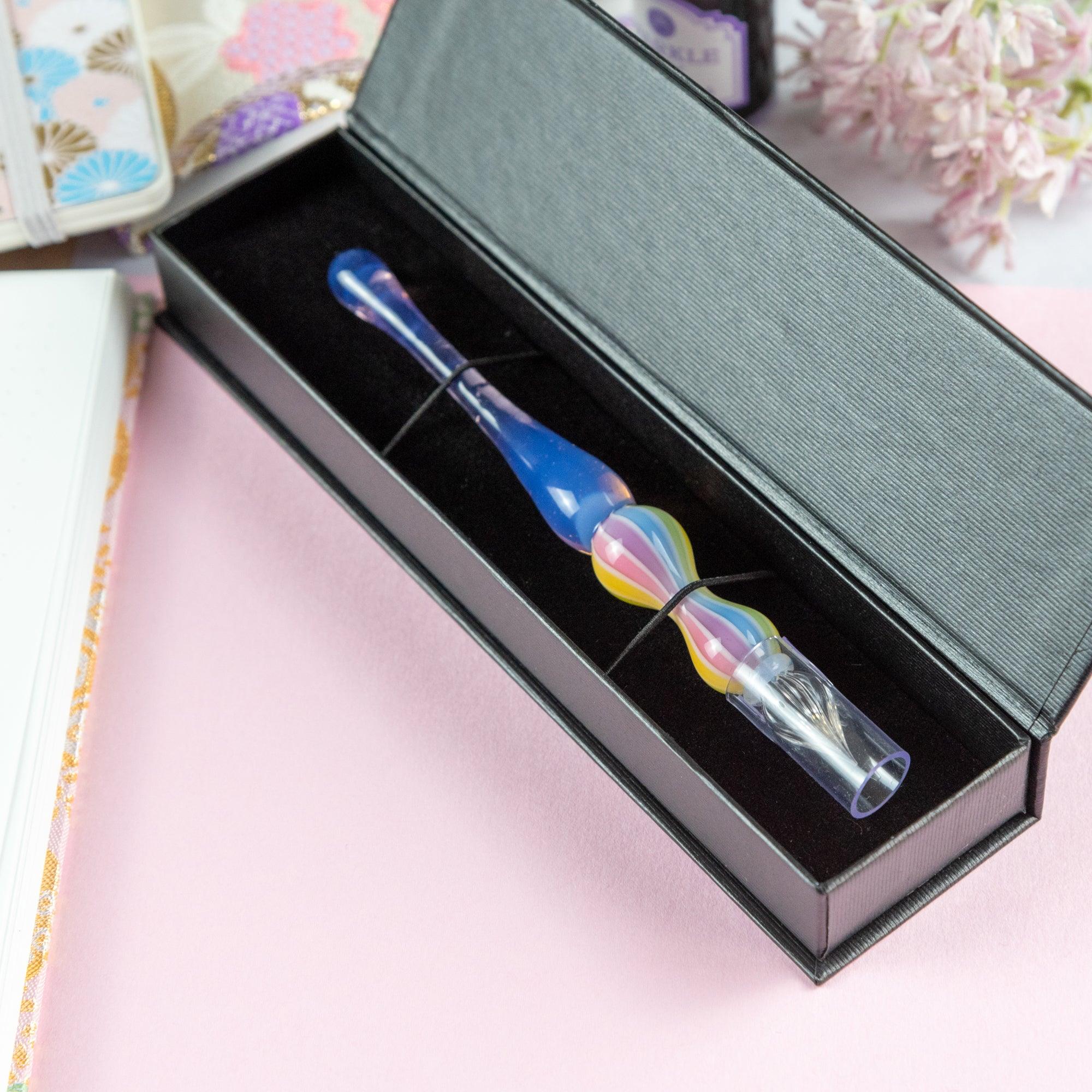 Pastel purple glass pen with rainbow-striped handle held while writing in a notebook. Ideal for Japanese stationery and glass pen fans.