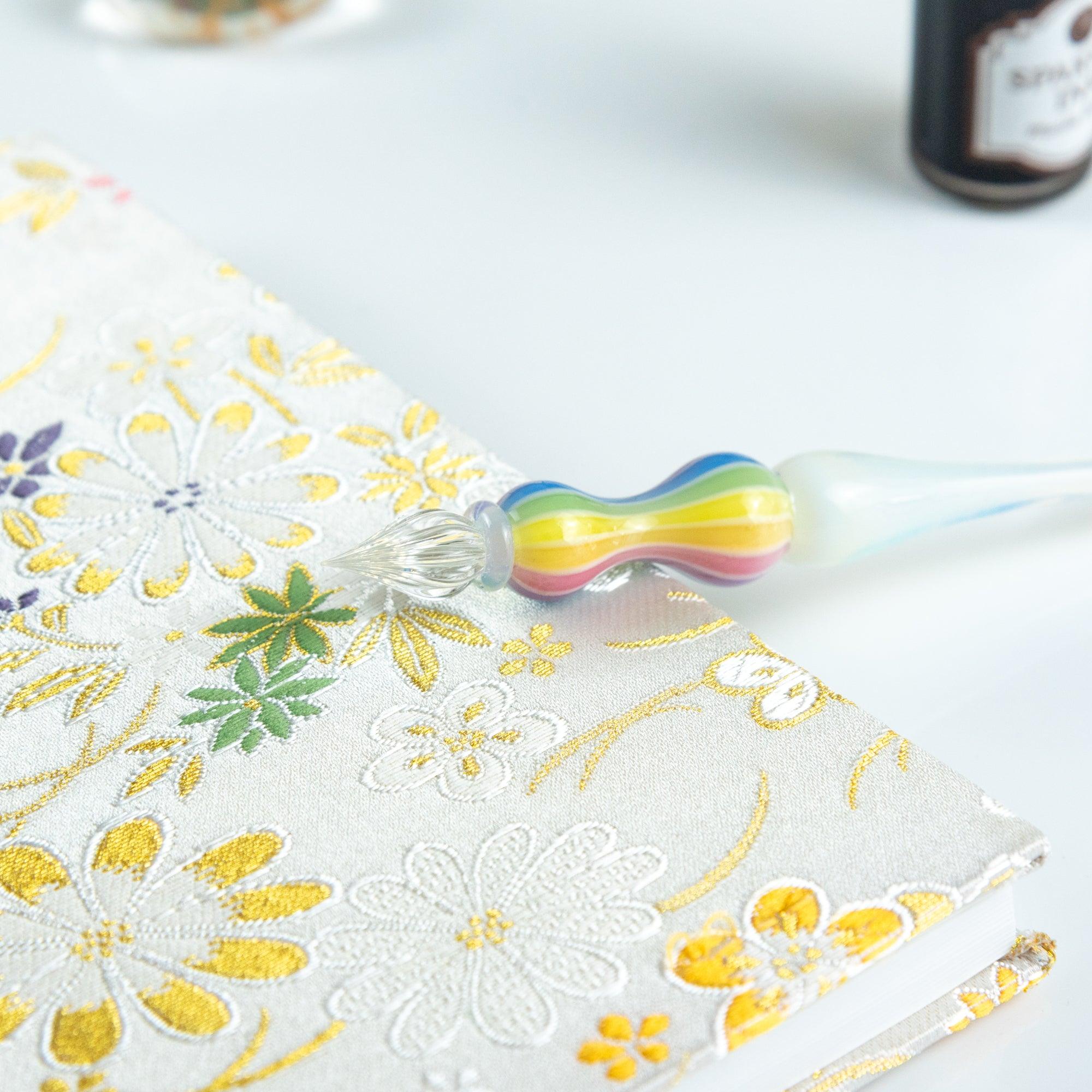 Glass pen with rainbow-colored handle resting on a floral-patterned notebook. Perfect for Japanese stationery enthusiasts.