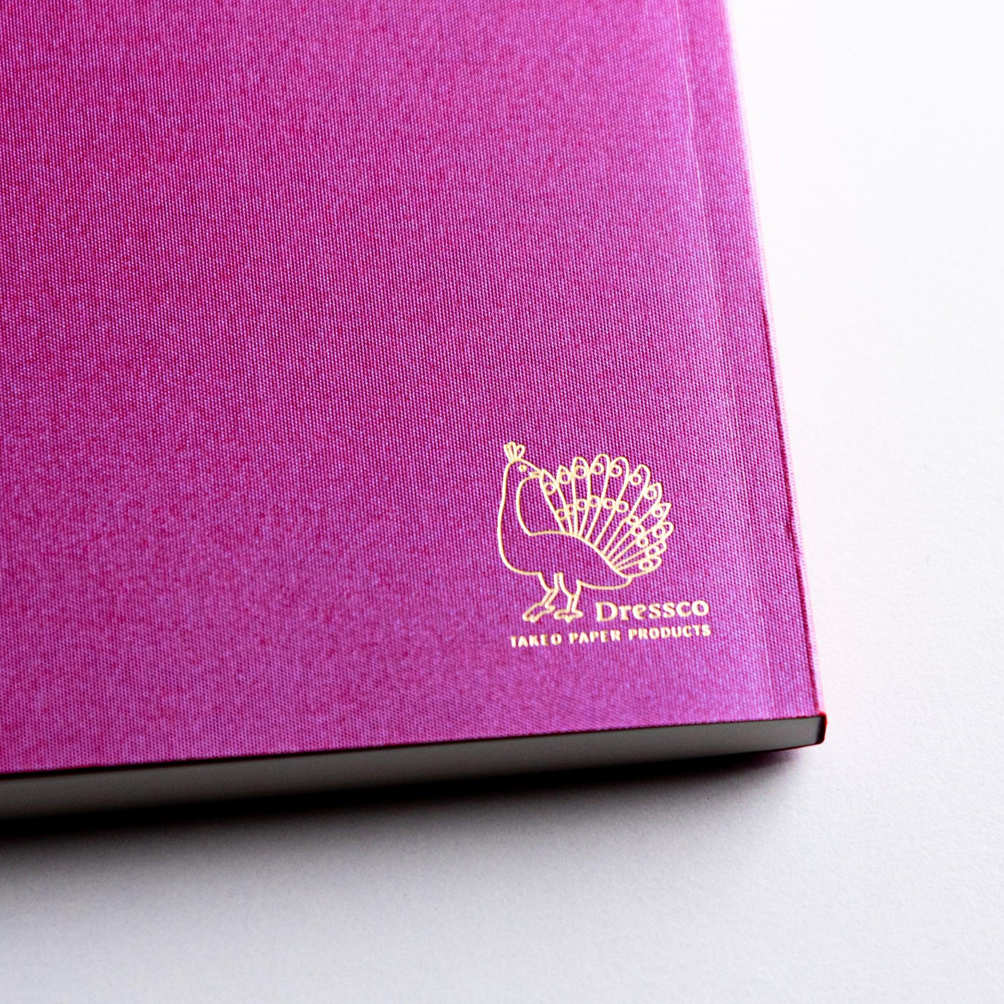 Close-up of gold Dressco peacock logo on textured pink cover of the Onion Skin Japanese Notebook A6.