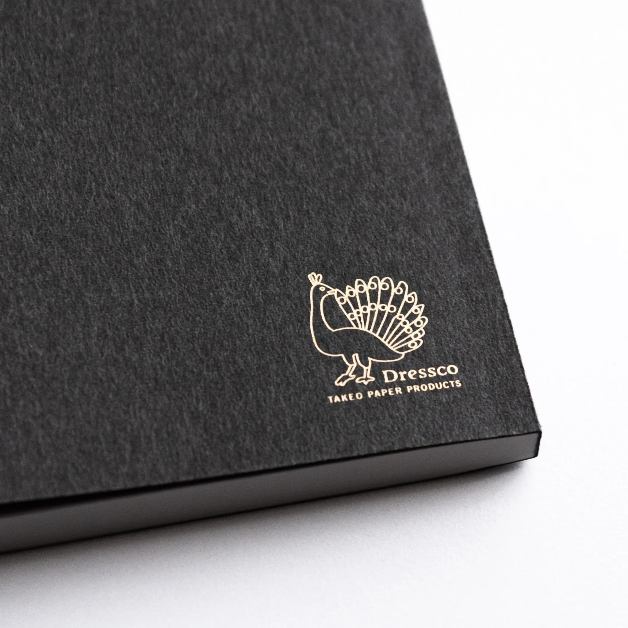 Close-up of gold Dressco peacock logo on textured gray cover of Japanese Notebook.