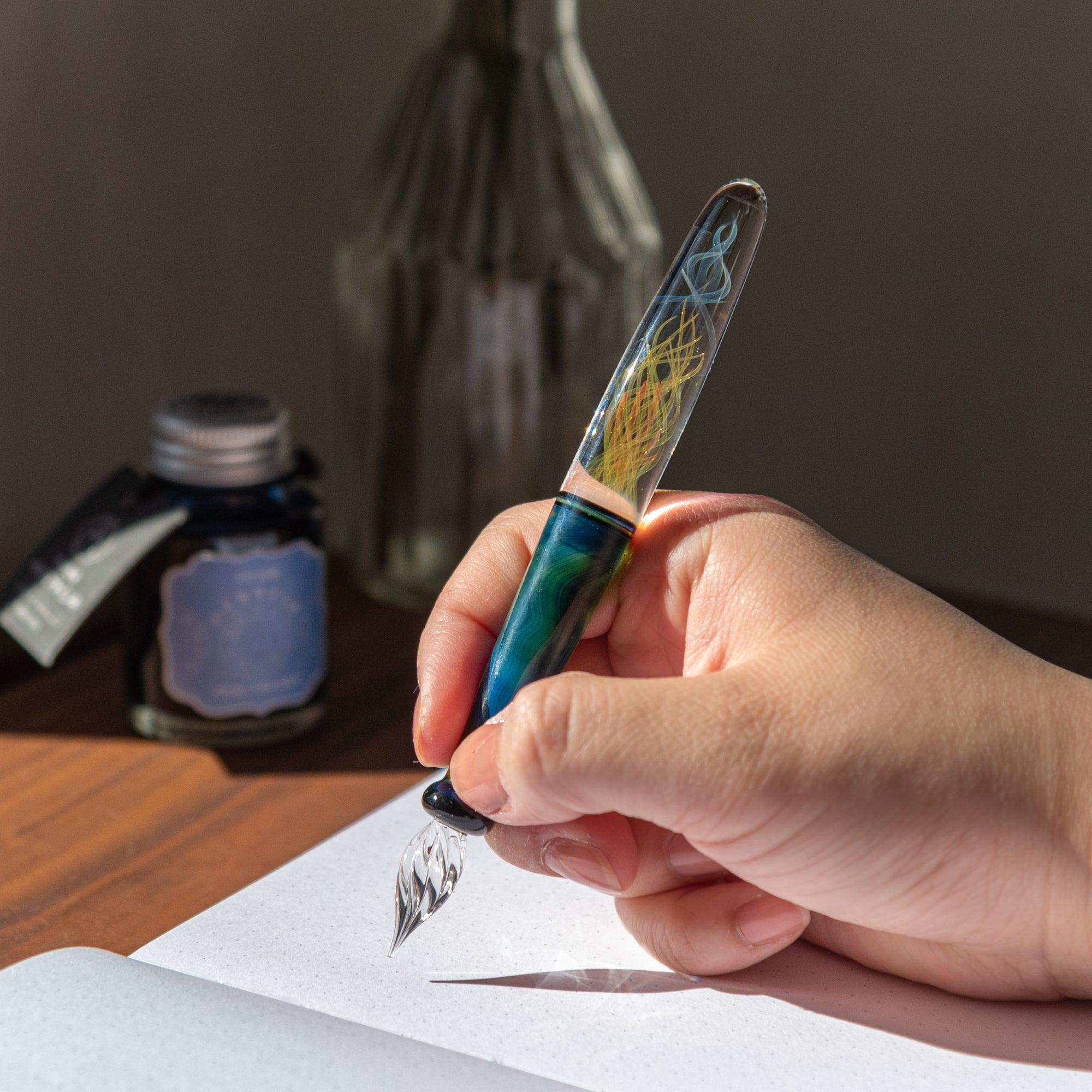 A hand holding the glass pen over a notebook. The colorful swirls inside the pen glow beautifully against the natural light in the background.