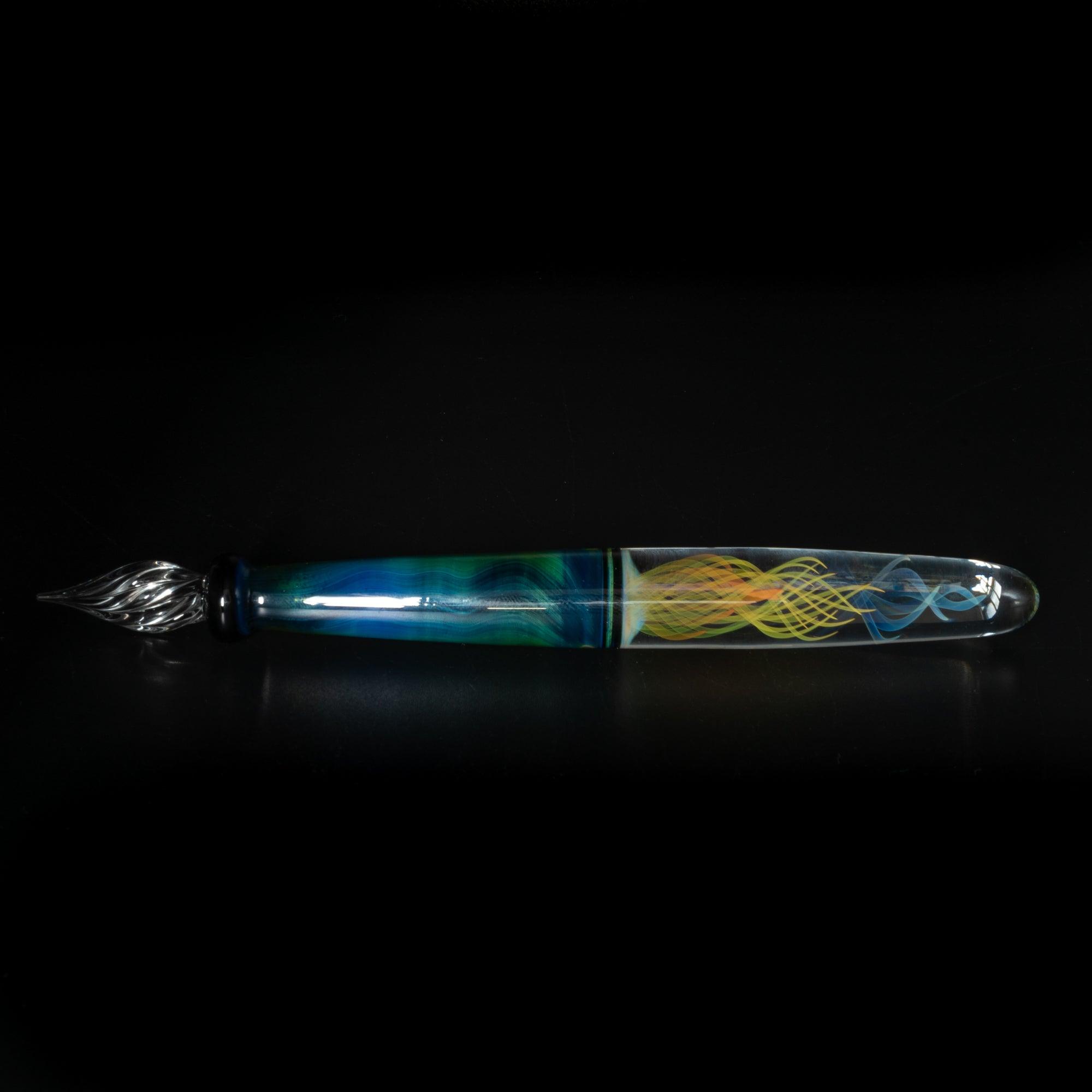 A glass pen with blue and green swirls, resting on a black surface. The clear nib transitions smoothly from the colorful body to the fine glass tip.