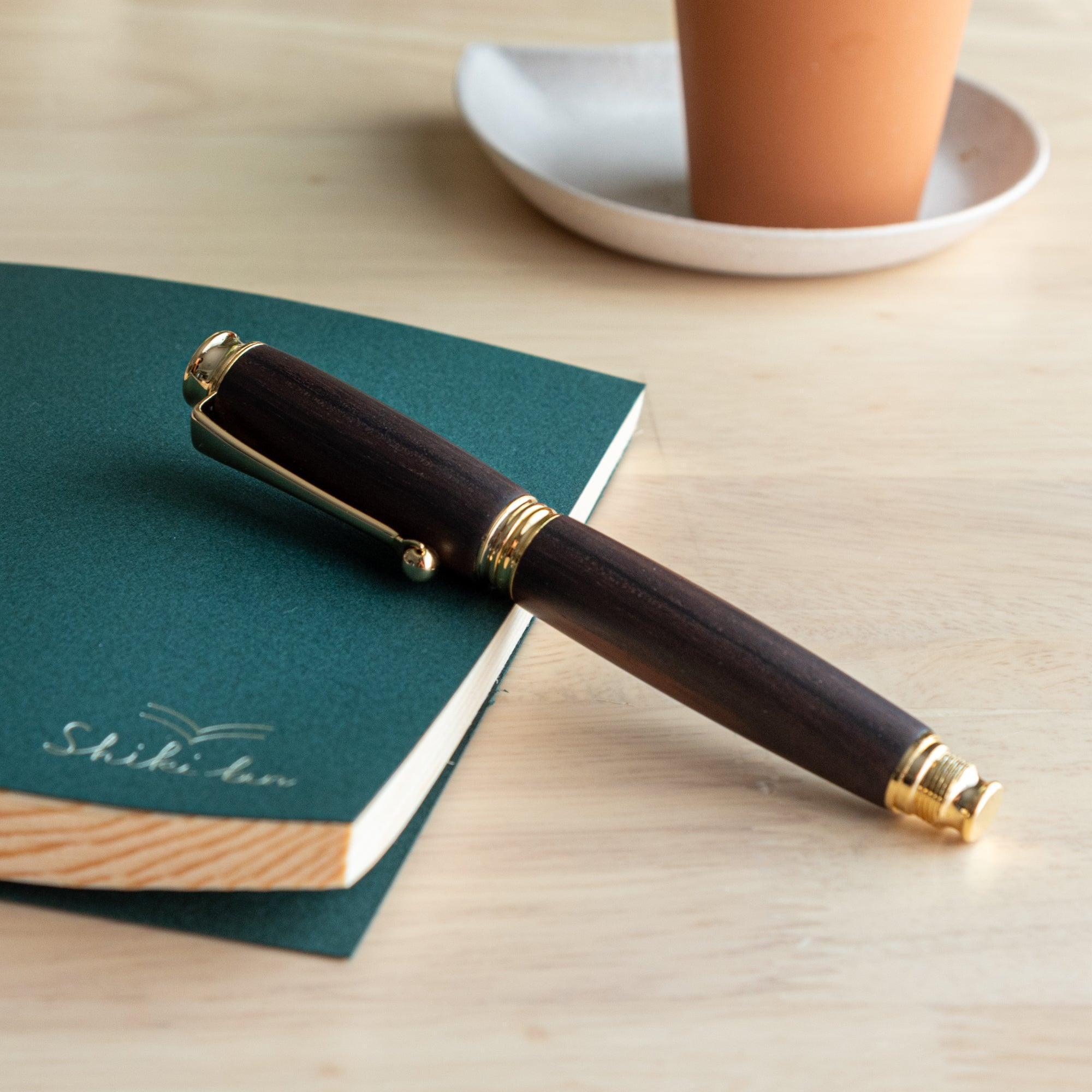 A Japanese ebony wood fountain pen with gold details resting on a green notebook, showcasing a refined design on a wooden desk.