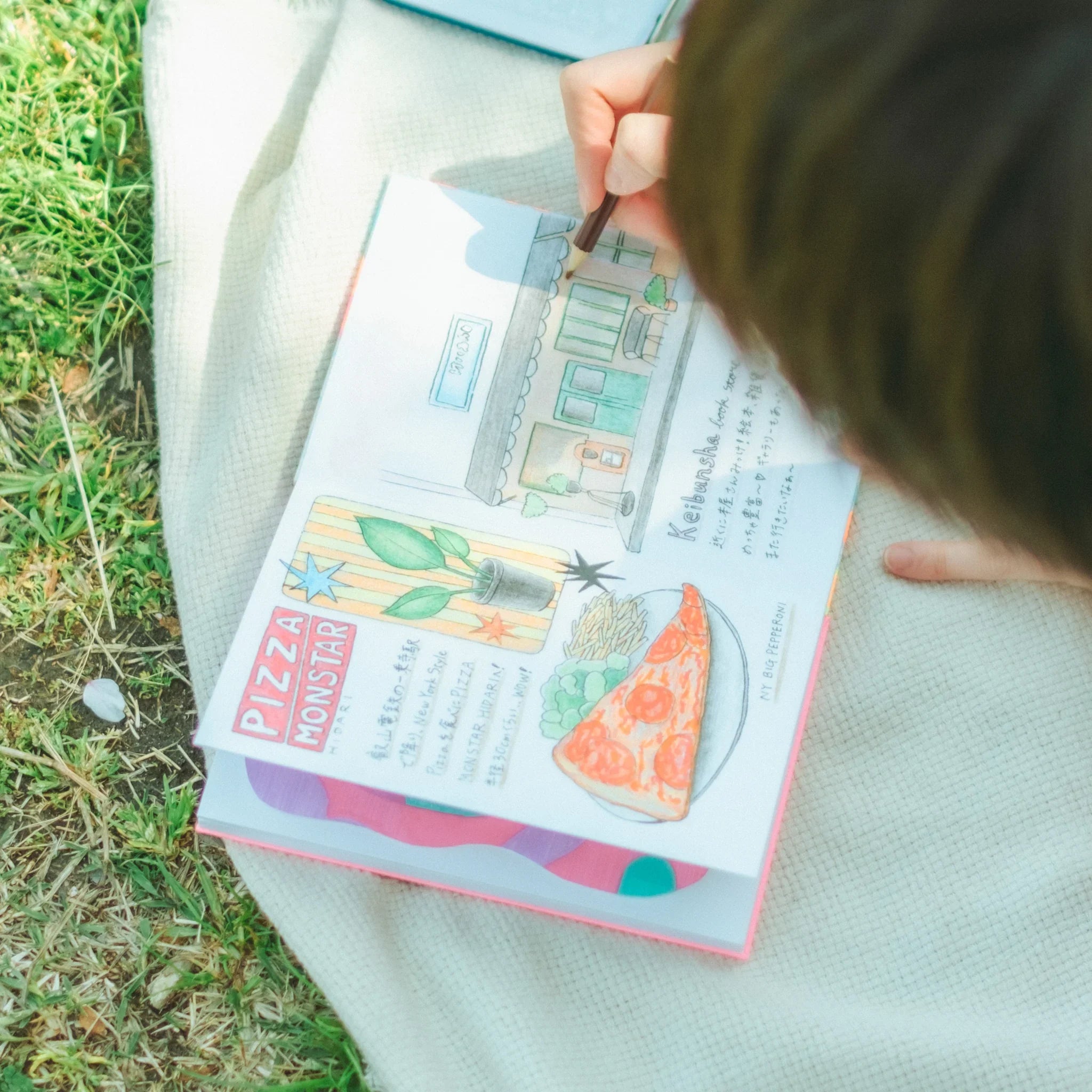 Person drawing colored pencil sketches of a pizza slice and shop front in a goshuincho notebook on a blanket outdoors
