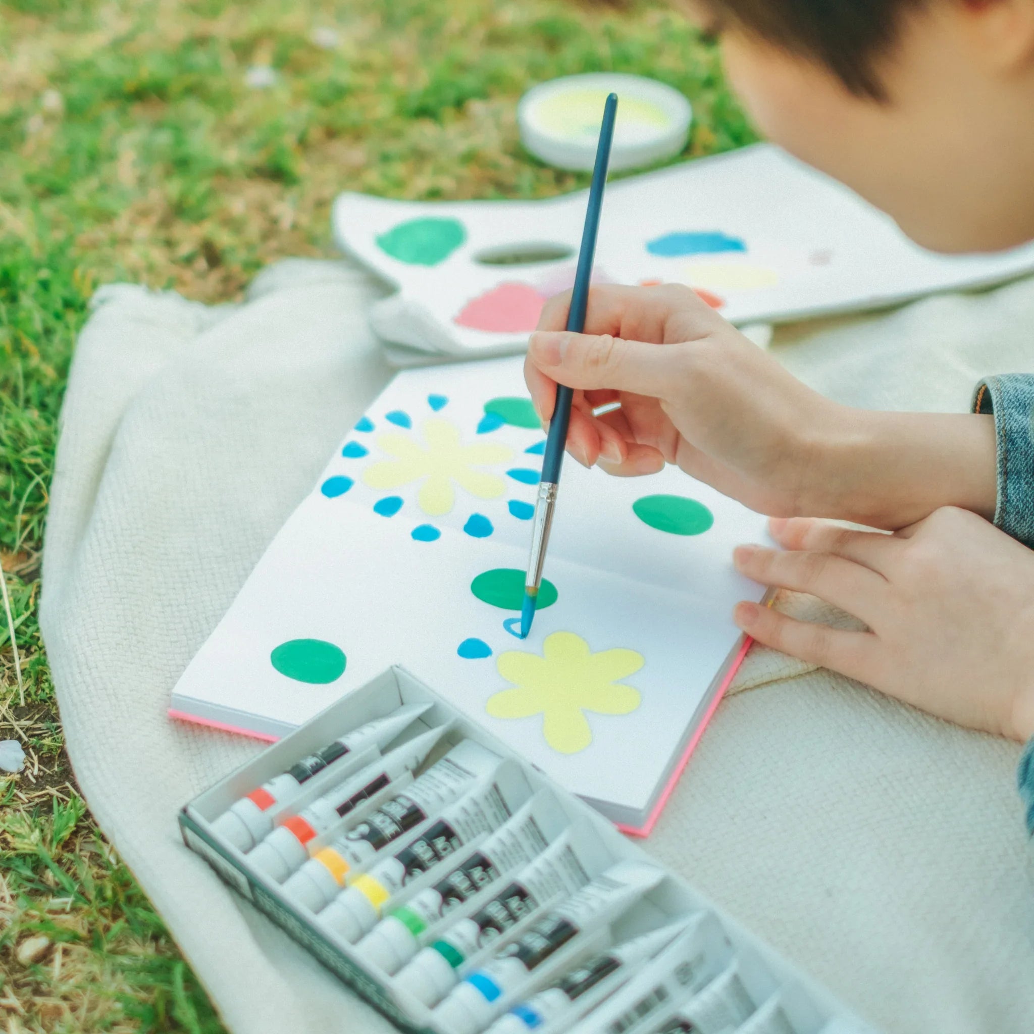 Person painting yellow and green flower shapes with a brush in an open goshuincho notebook, paint tubes and palette nearby