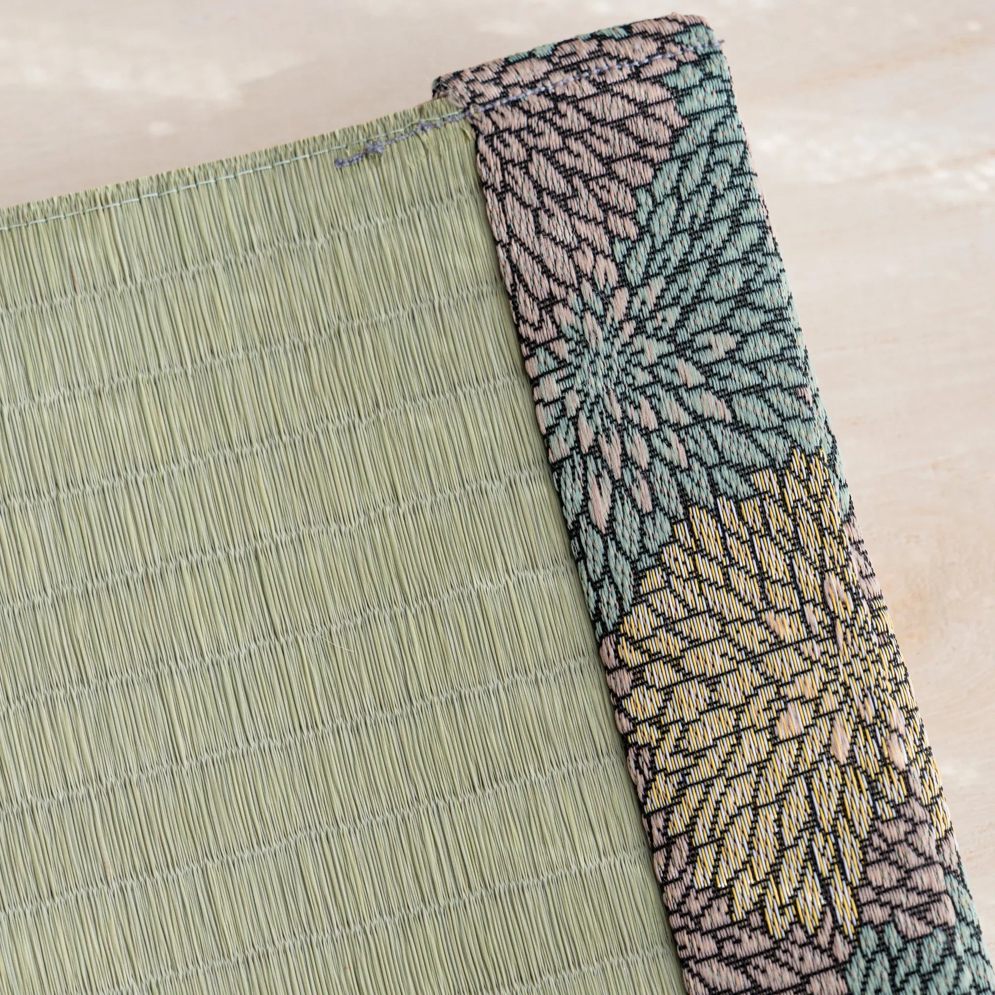 Angled close-up of the chrysanthemum brocade band beside woven green rush straw at the tatami book cover corner