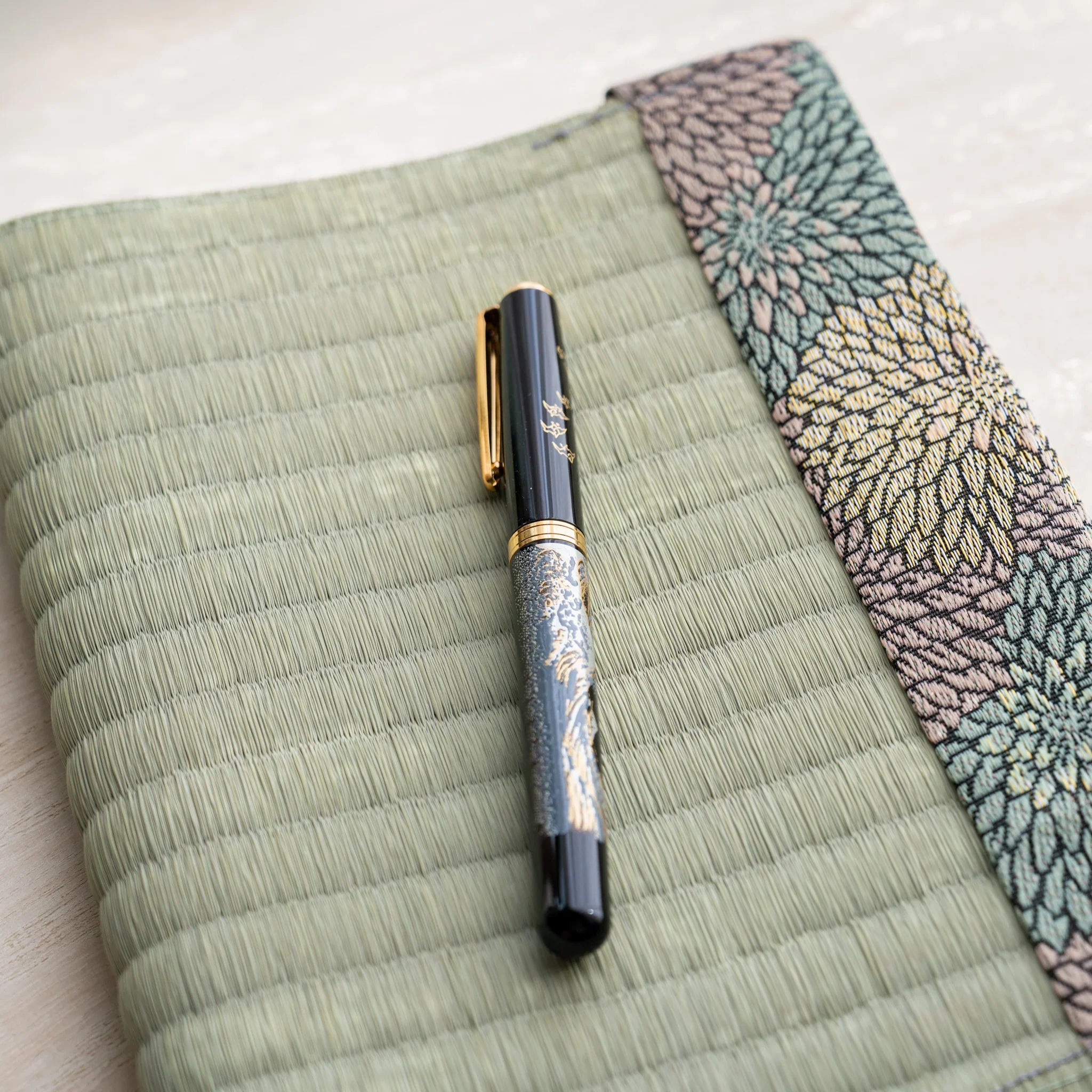 Close-up of a black fountain pen on the woven green rush straw cover beside the chrysanthemum patterned brocade band