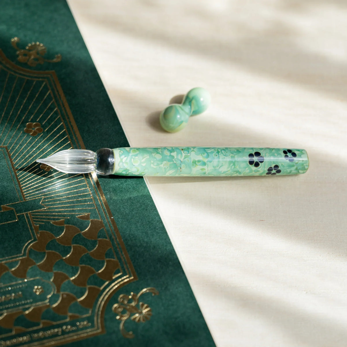 Emerald Flower glass dip pen laid across a green notebook with gold foil printing, pen rest above