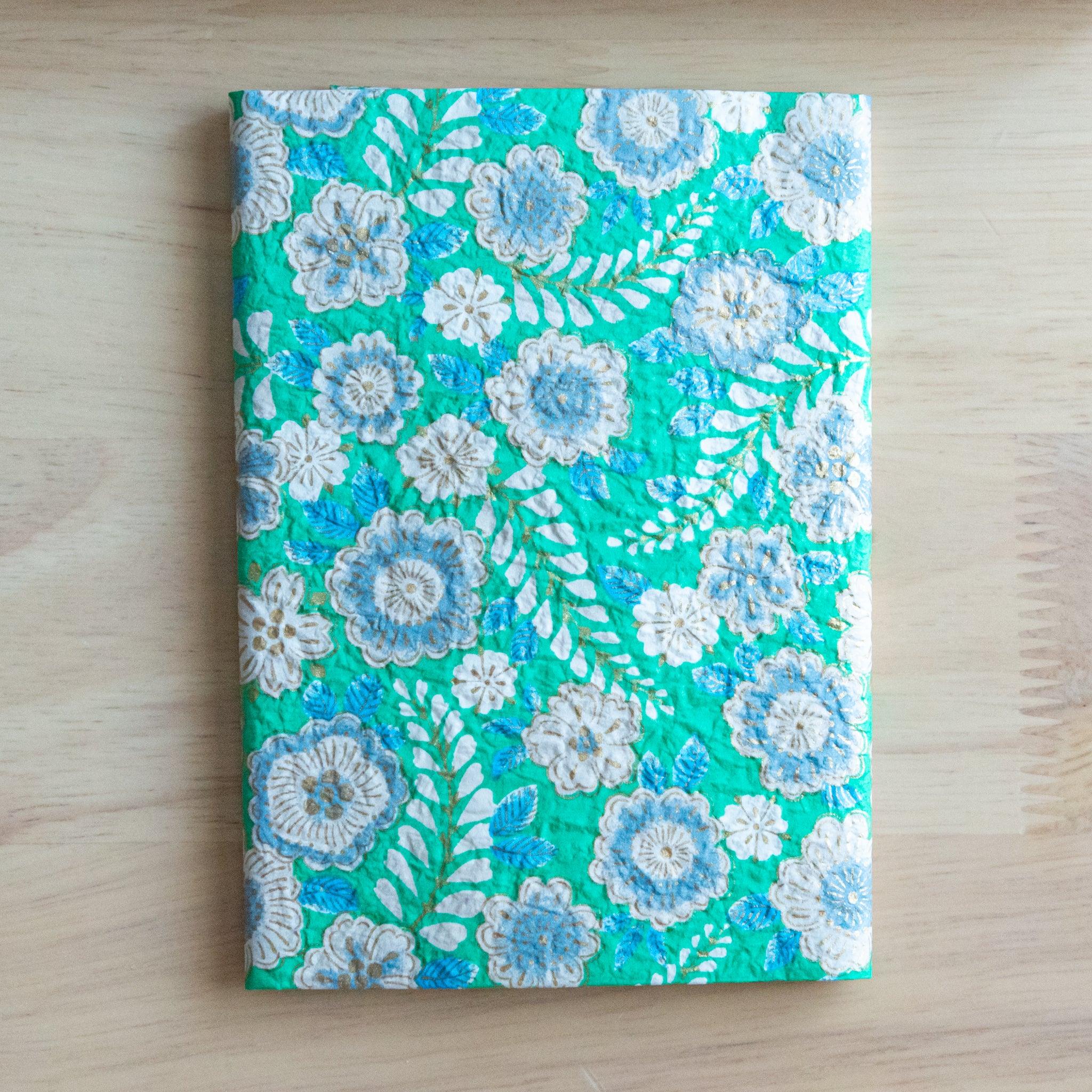 An open notebook cover with a green floral pattern featuring white and blue flowers, revealing its inner white lining.