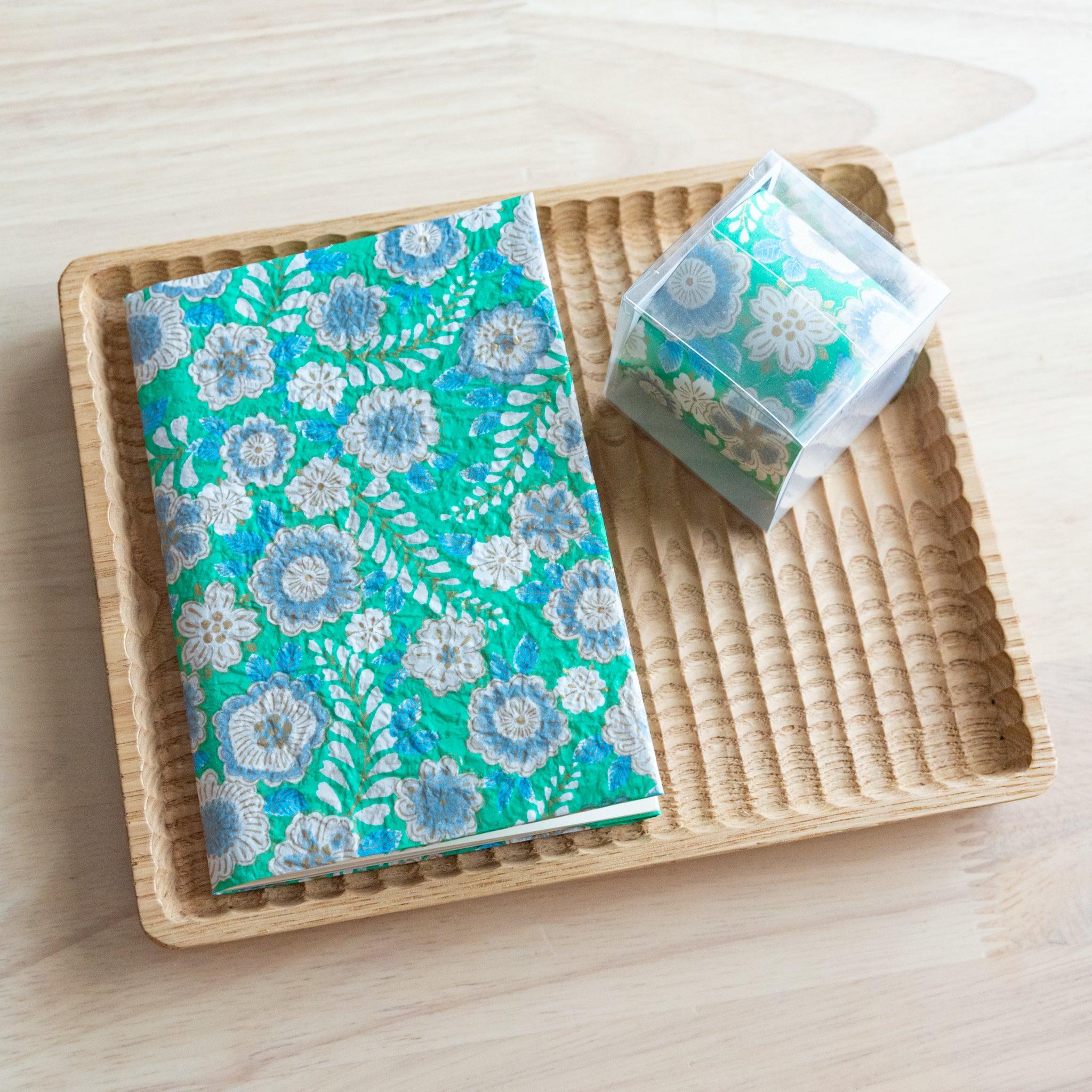 A notebook with a green floral pattern, decorated with white and blue flowers, placed on a light wooden surface.