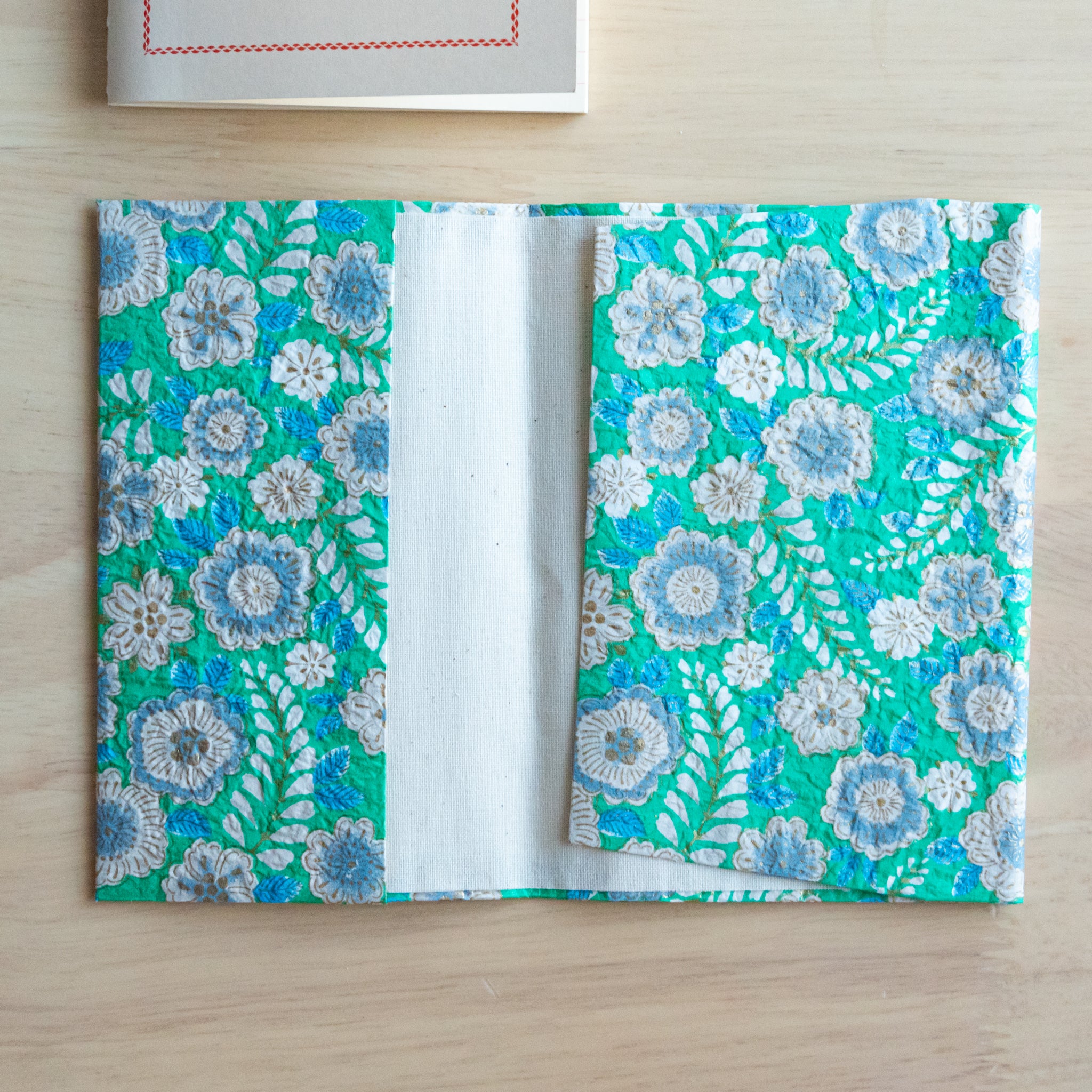 A green floral patterned notebook with white and blue flowers, placed on a wooden tray alongside matching washi tape.