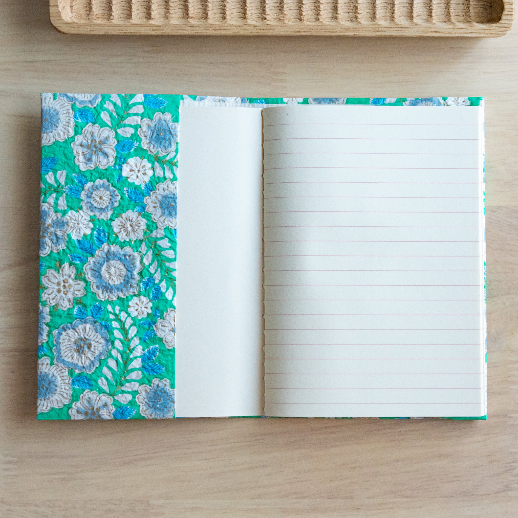 An open notebook with green floral patterned covers, showing one blank page and one lined page on a wooden surface.