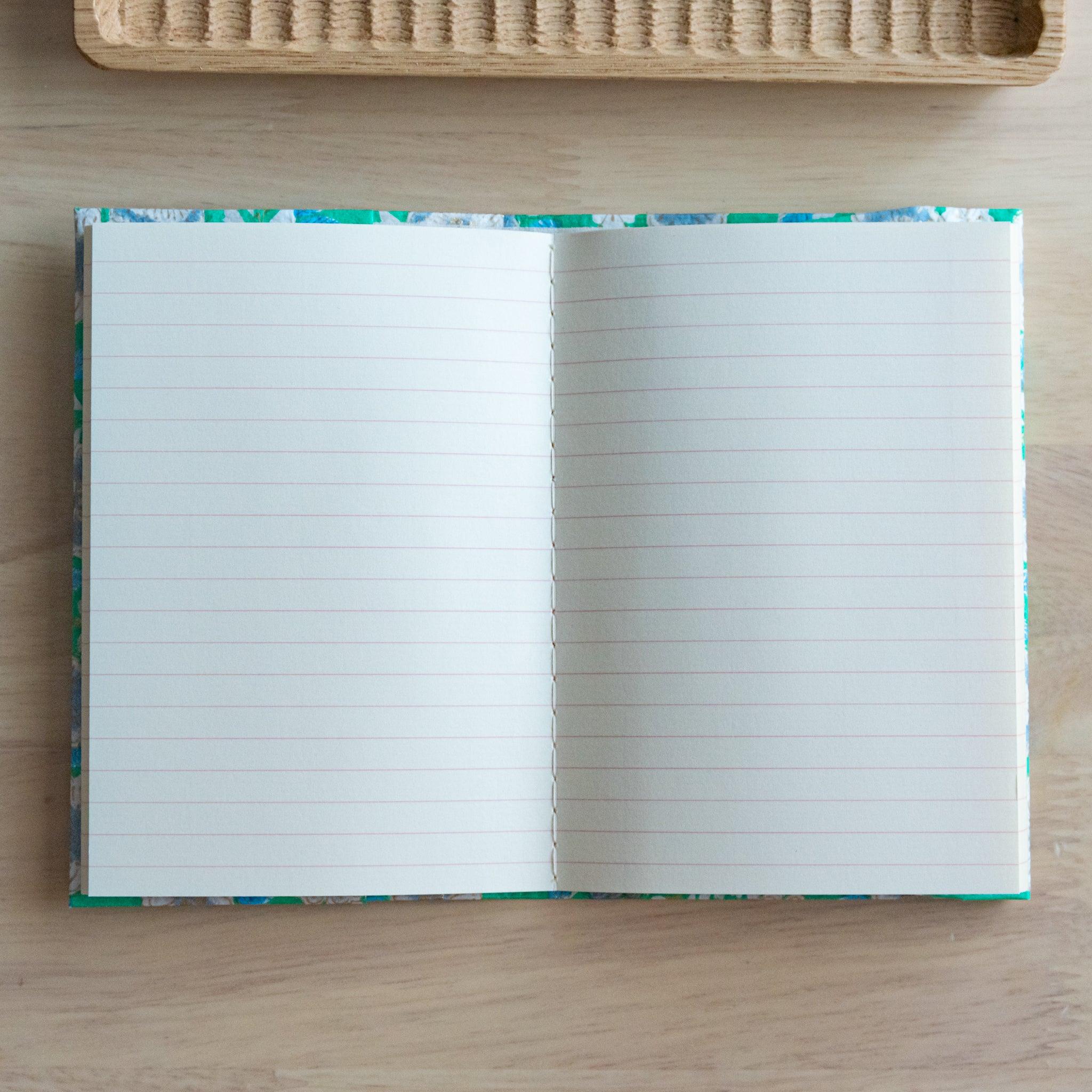 An open notebook with green floral patterned covers, fully revealing two lined pages, placed on a light wooden surface.