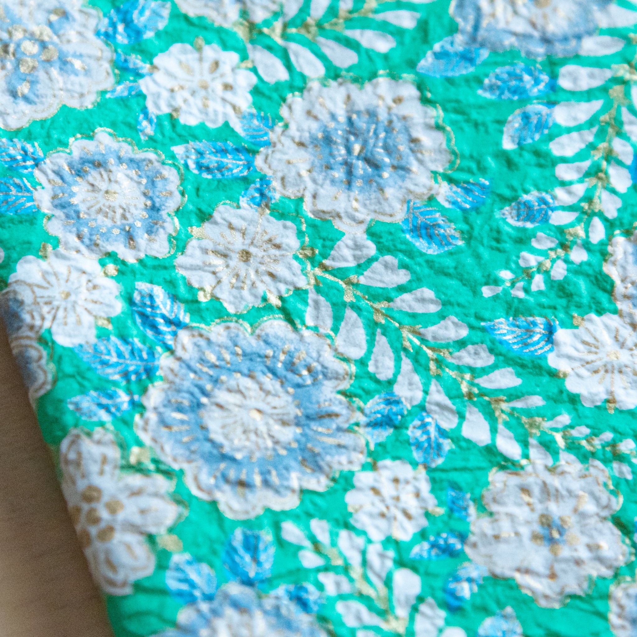 A close-up of a green floral pattern featuring white and blue flowers with gold accents, showing detailed textures on the paper.