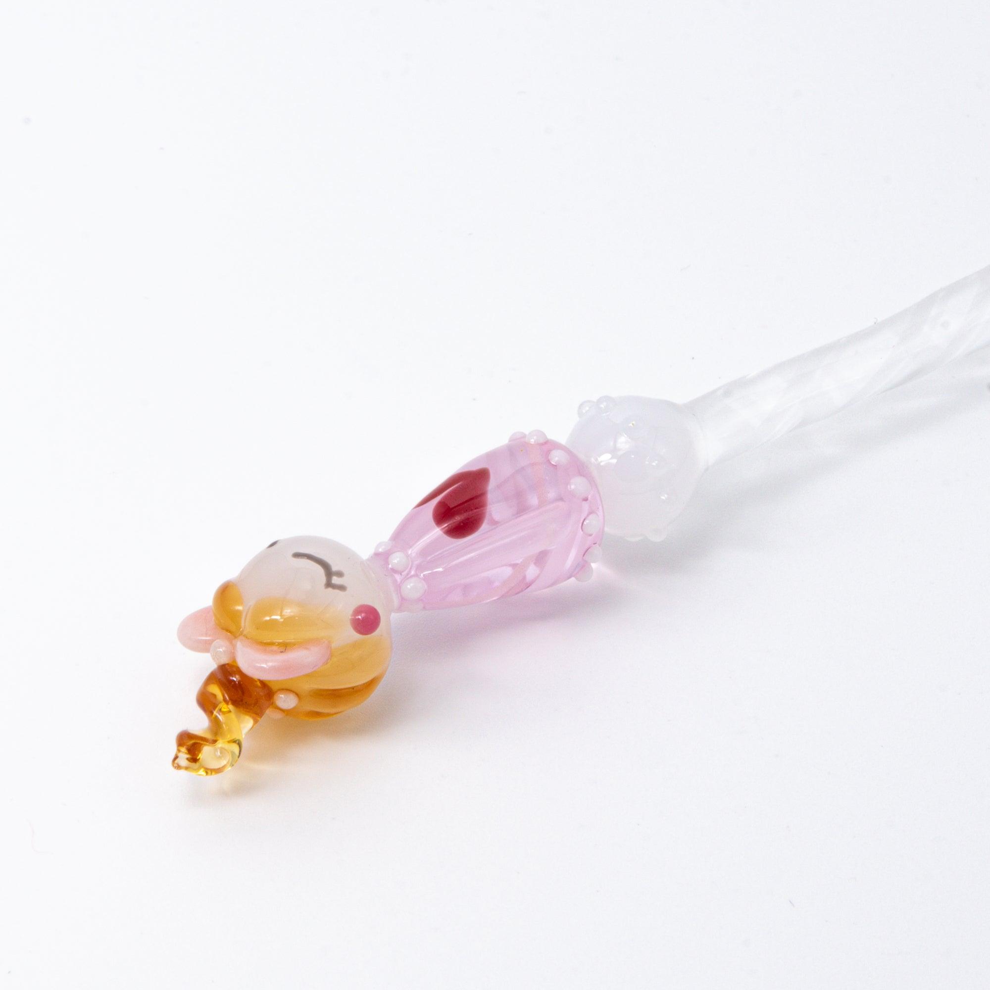 A top view of a Lovery-chan glass pen with a heart design, featuring a cute figure on top, dressed in pink with hearts along the pen shaft.