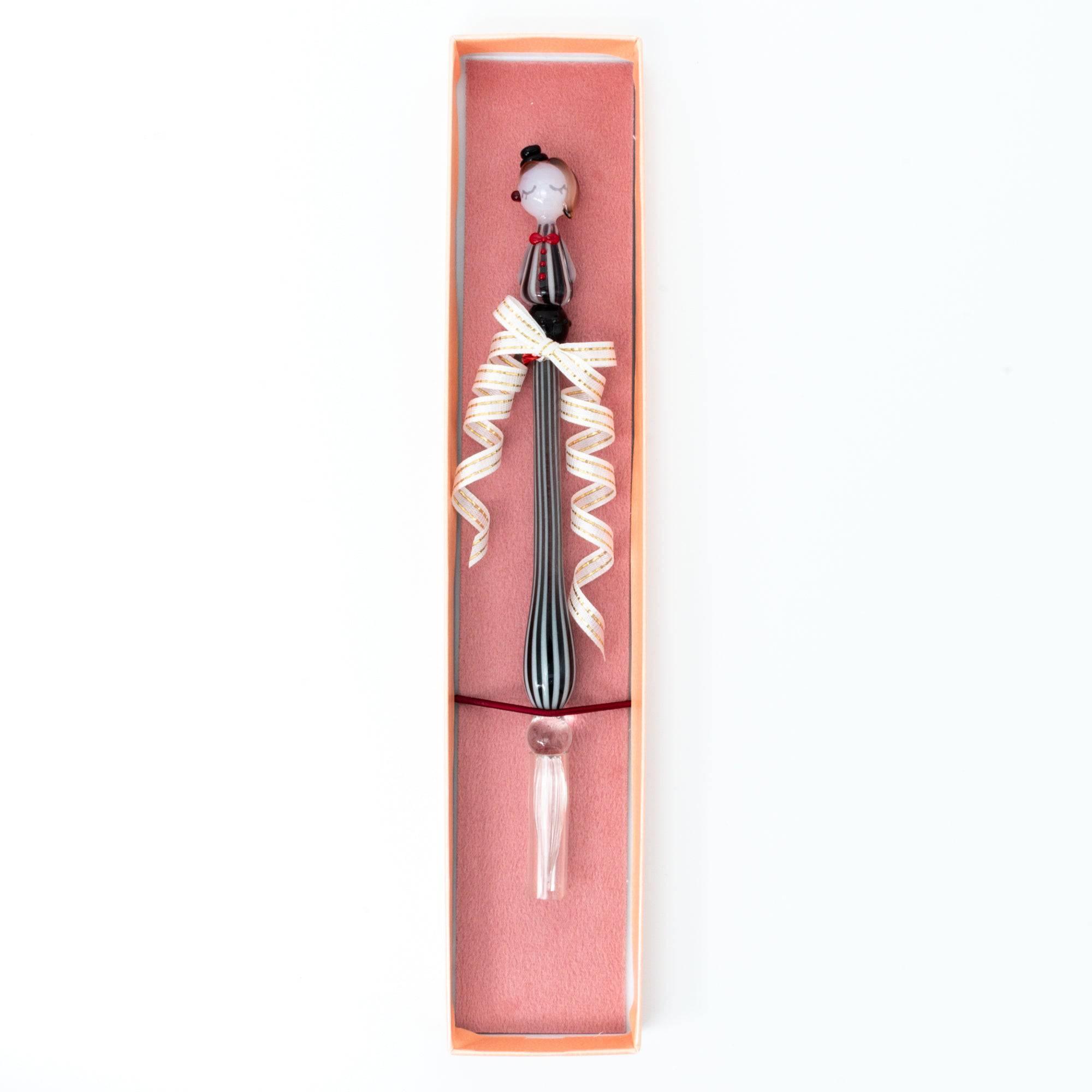 A striped glass dip pen with a whimsical lady figurine top, neatly displayed in a pink gift box tied with a ribbon