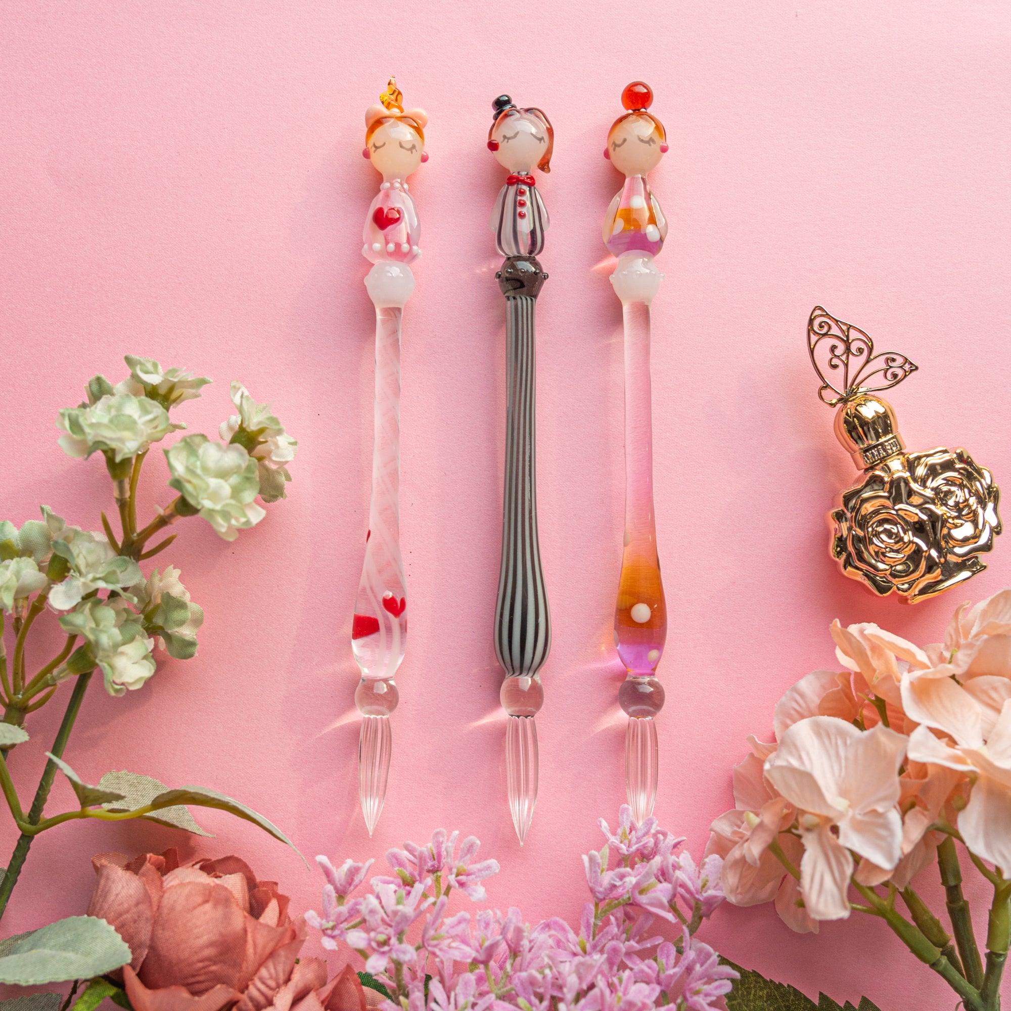 Three glass dip pens with whimsical lady figurine tops, surrounded by flowers and a decorative perfume bottle on a pink background.