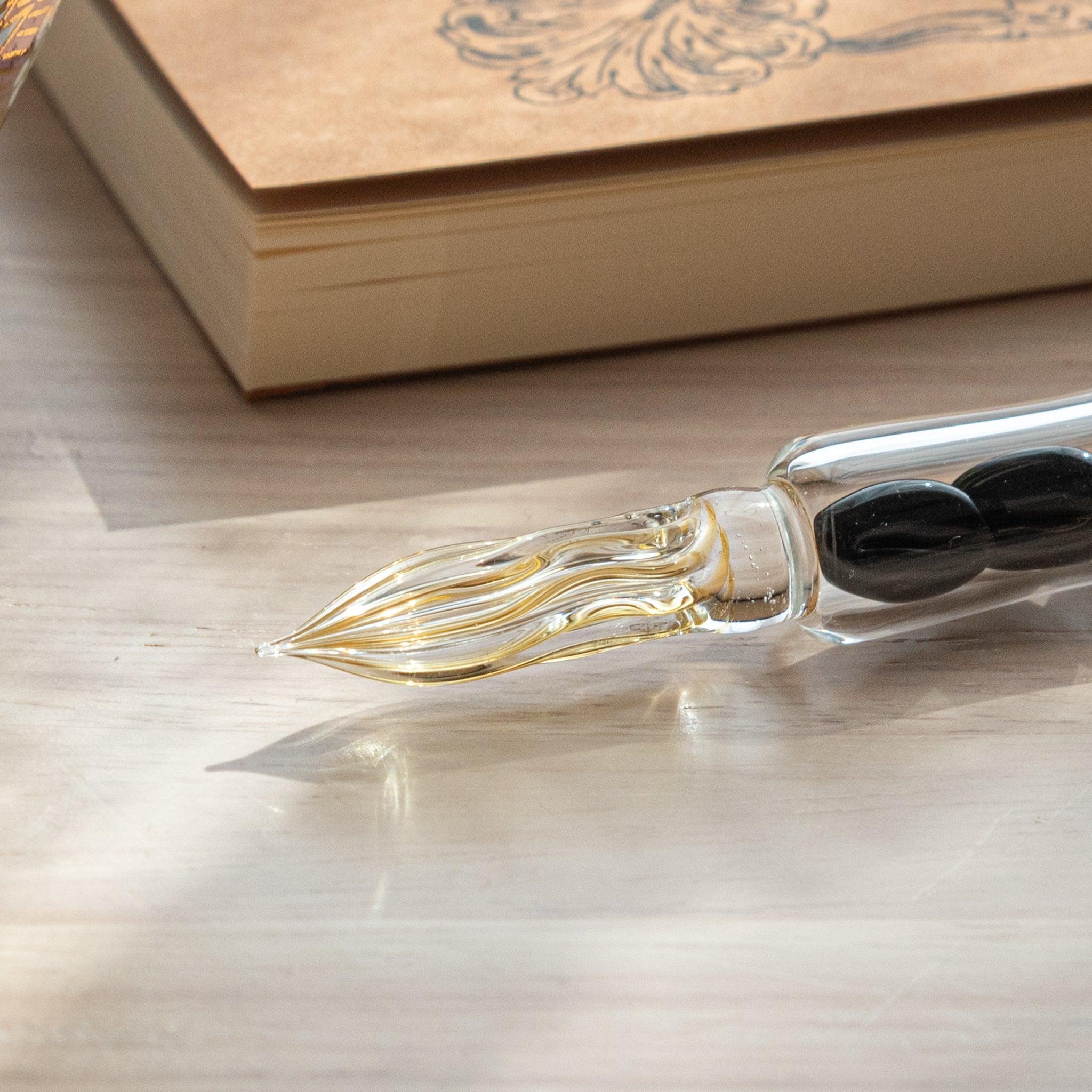 A glass dip pen with coffee bean-shaped glass pieces inside its shaft and a coffee-stained colored nib rests on a wooden surface near coffee beans and a plain notebook.