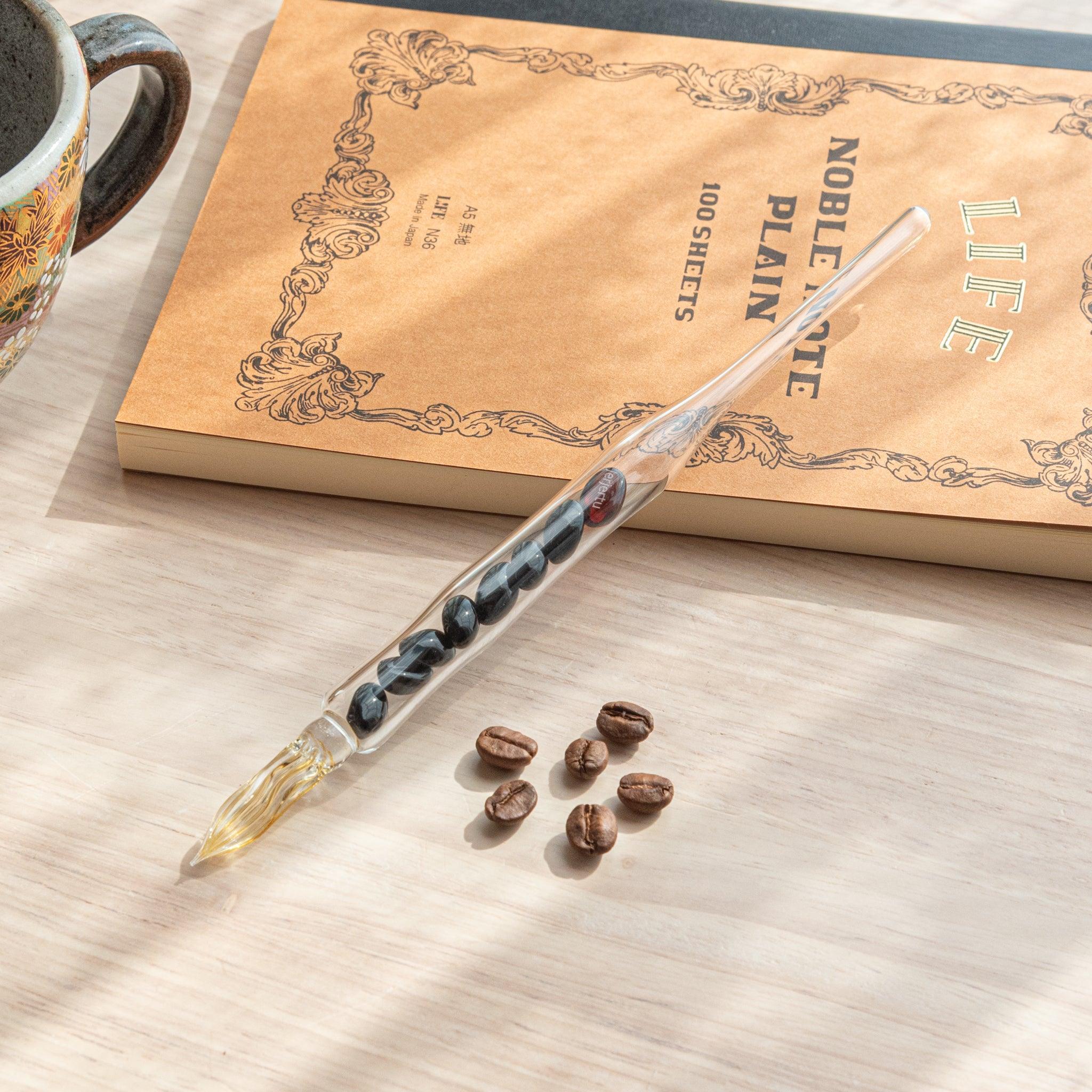 A glass dip pen with coffee bean-shaped glass pieces inside its shaft and a coffee-stained colored nib rests on a wooden surface near coffee beans and a plain notebook.